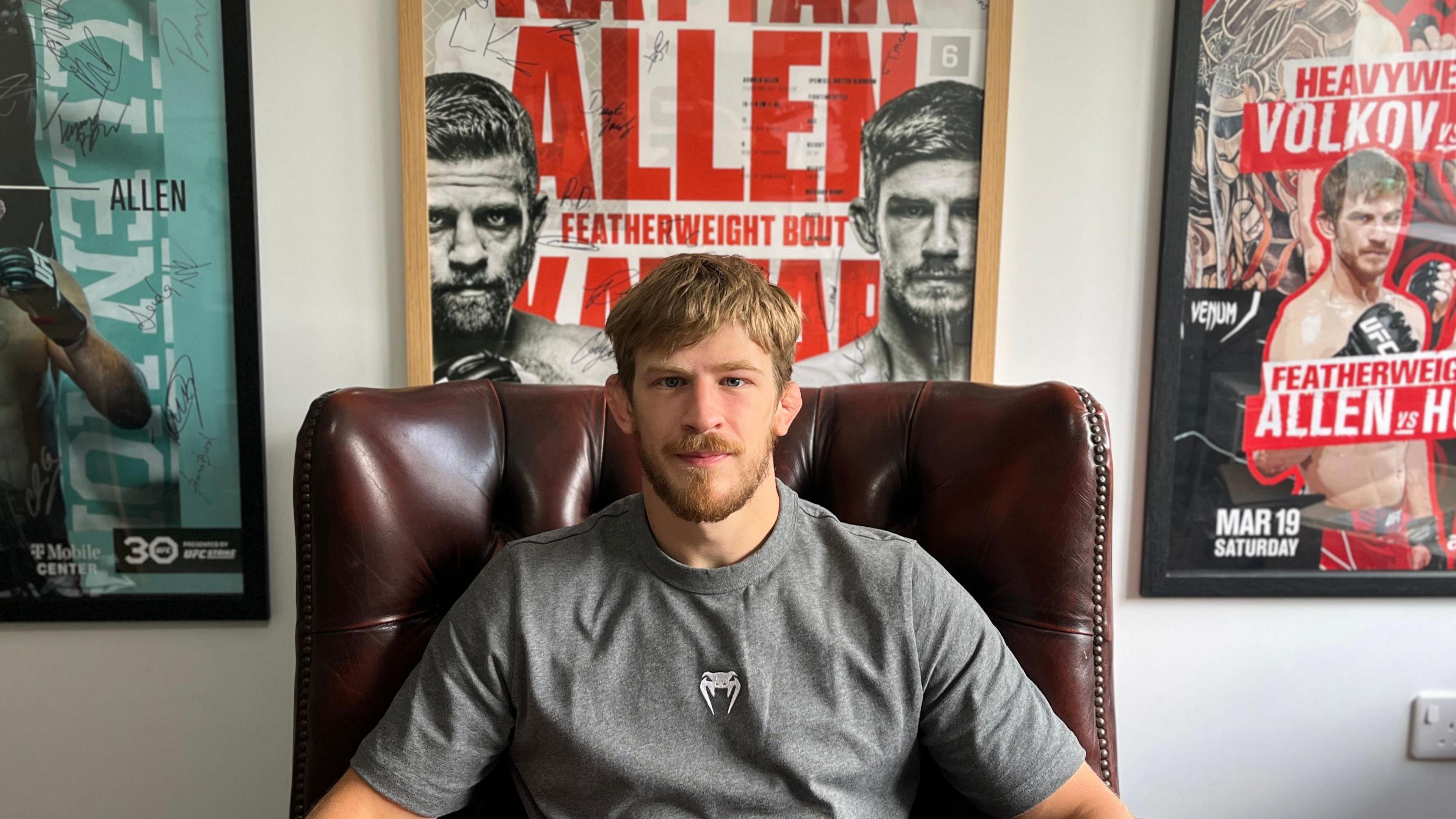 A picture of Arnold Allen. He is sitting in a brown leather chair facing the camera. He has blond hair and a blond beard. He is wearing a grey t-shirt. Posters from his fights are placed on the wall behind him.