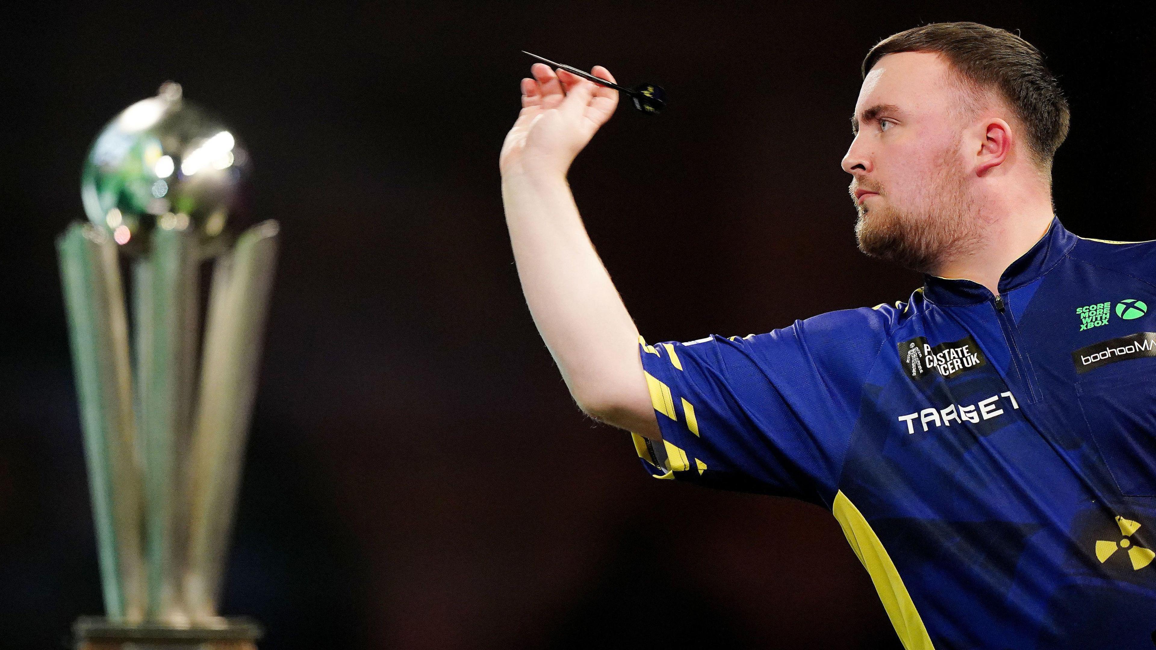 Luke Littler throwing a dart with the world championship trophy in the background