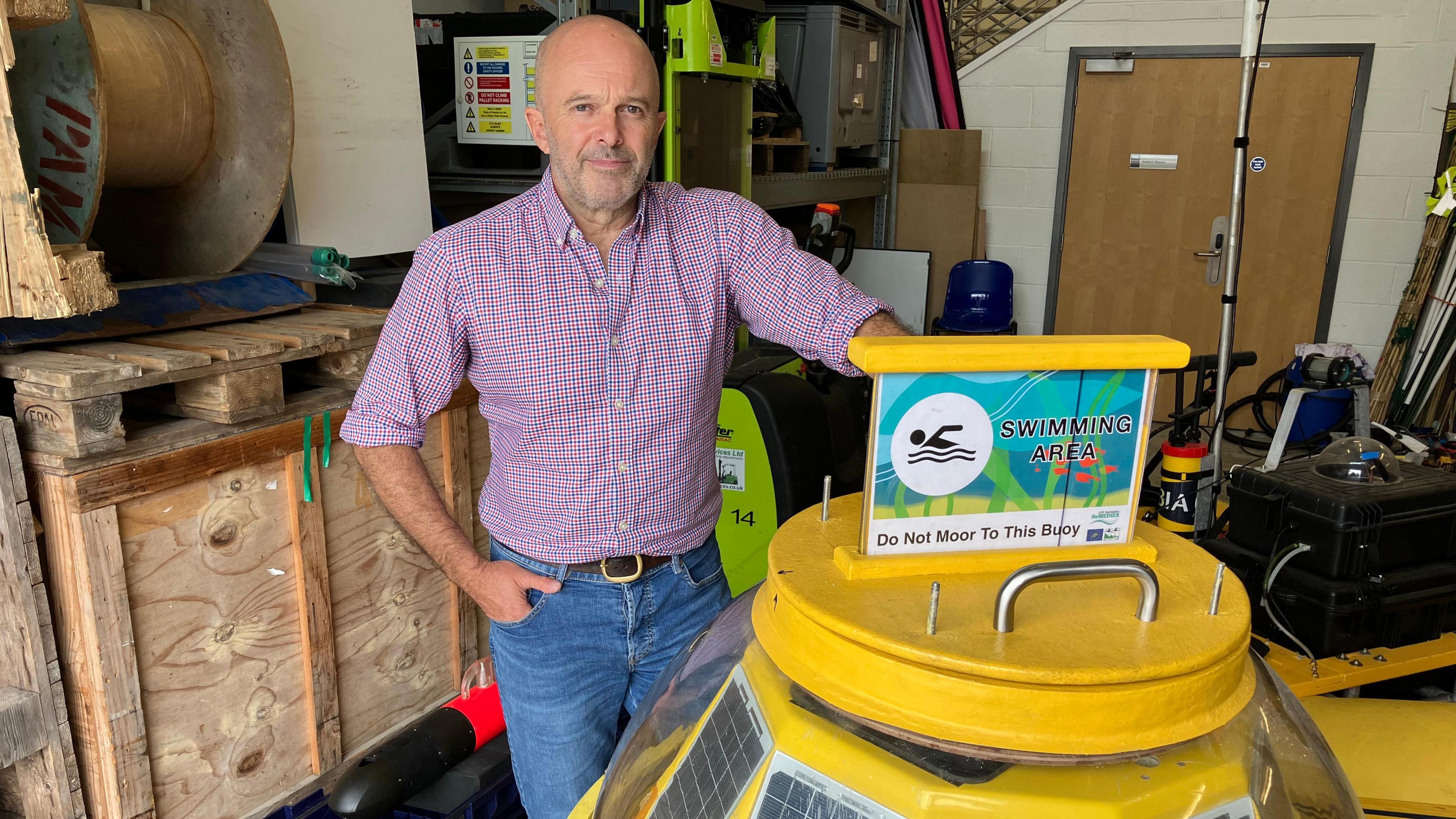 A man stands in a large warehouse - a marine science workshop - next to large yellow buoy. It is waist height and is covered in solar panels.
