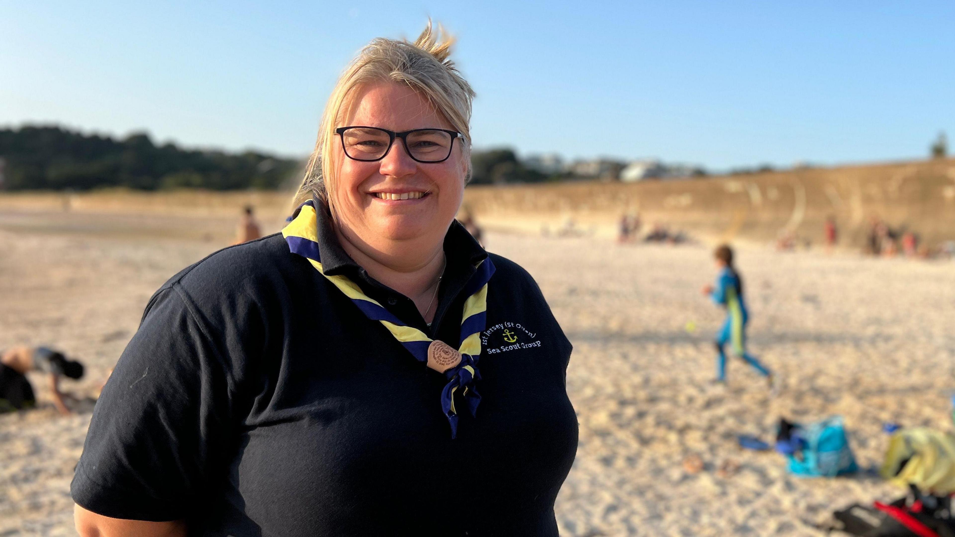 Sam, who is wearing a navy blue shirt and a yellow and blue neckerchief, stands on a sandy beach. In the background, other people are sitting or walking on the beach, some carrying bags or wearing swimsuits. A cliff is visible in the distance under a clear blue sky.
