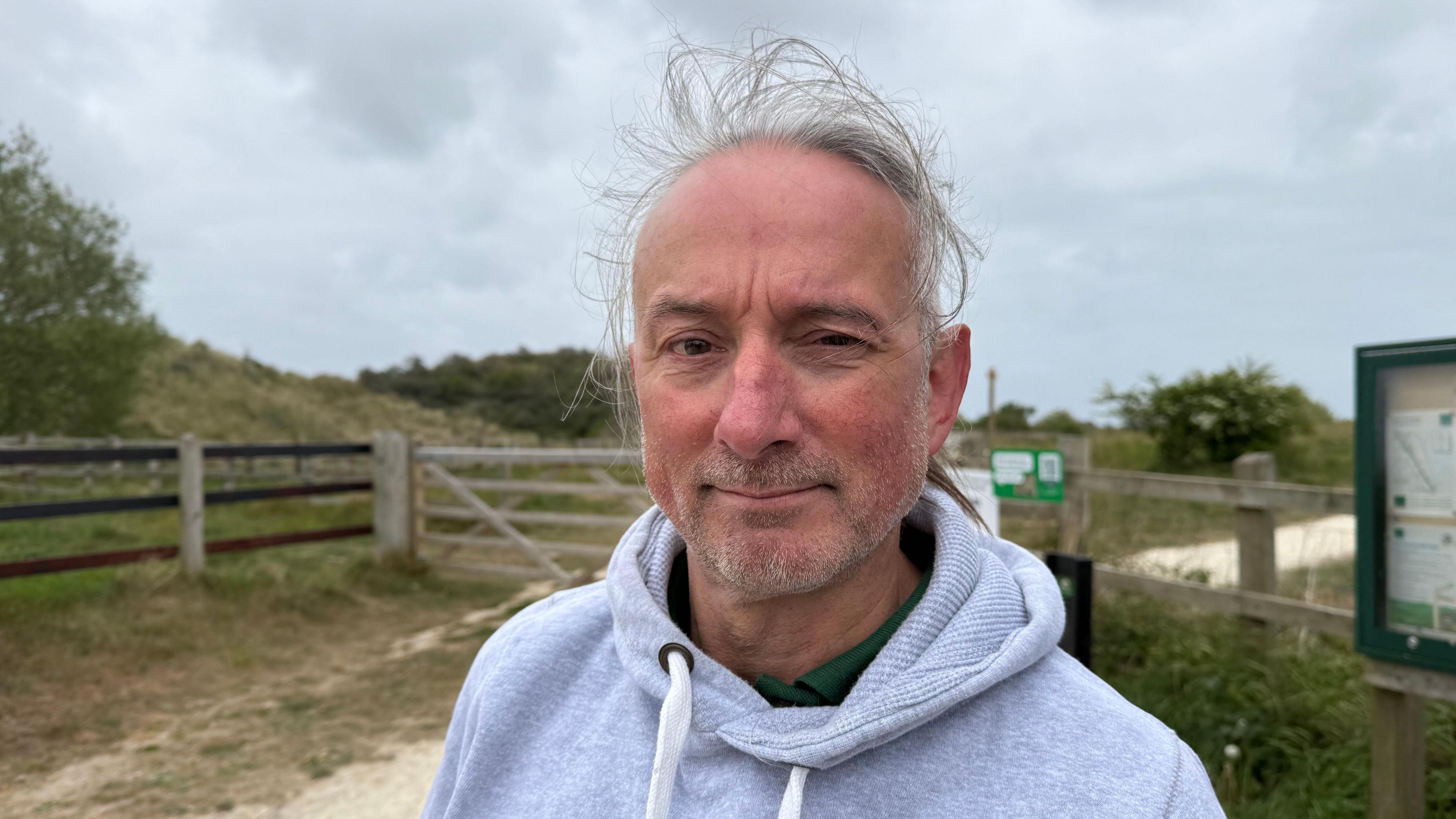 A man is at the centre of the photo. He's wearing a grey hoodie, and has grey hair that is tied up in a ponytail. He has a beard, and is smiling at the camera. In the background there is a gate, grass, plants and trees.