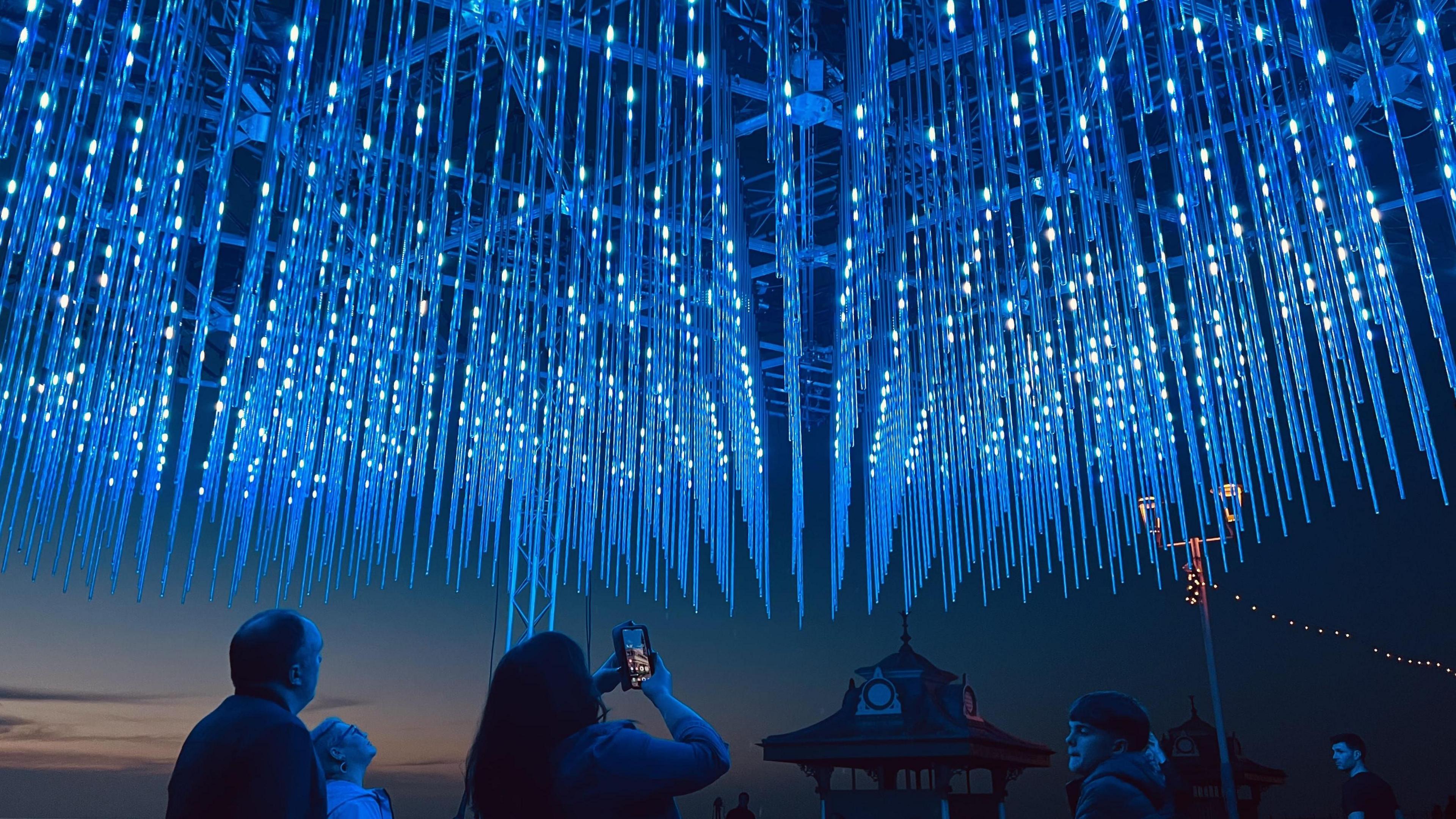 Many blue strobe like lights hanging from the ceiling. There are people on the ground looking up at the sky. A woman in the centre is taking a photo of the lights with her mobile phone.