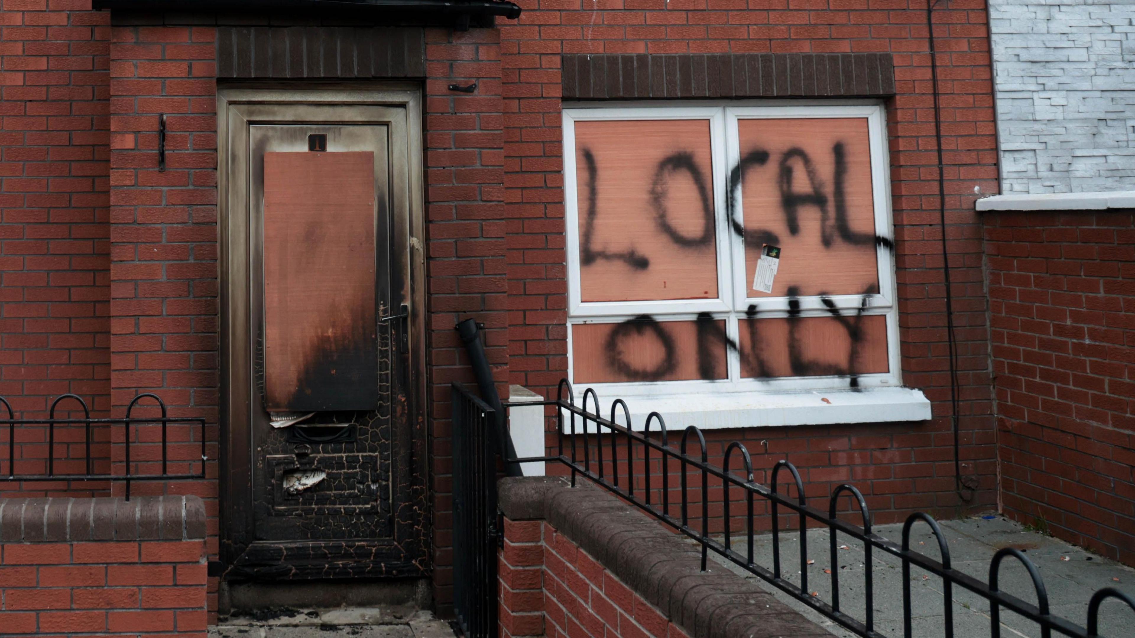A view of the vandalised house.  The red-brick terraced property has windows on either side of the front door on both its ground and upper floors.  The downstairs windows have been boarded up and the words "local only" have been sprayed on the right hand side window.  The white PVC front door has sustained smoke and fire damage. 