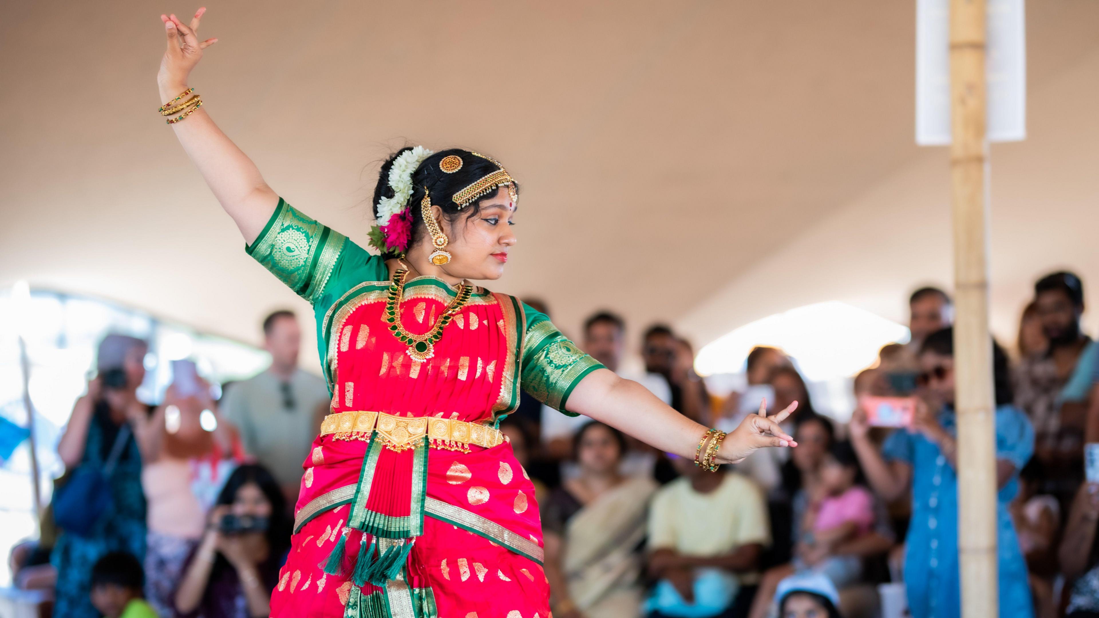 A woman in a sari is dancing with her arms extended. There is a crowd watching her in the background.