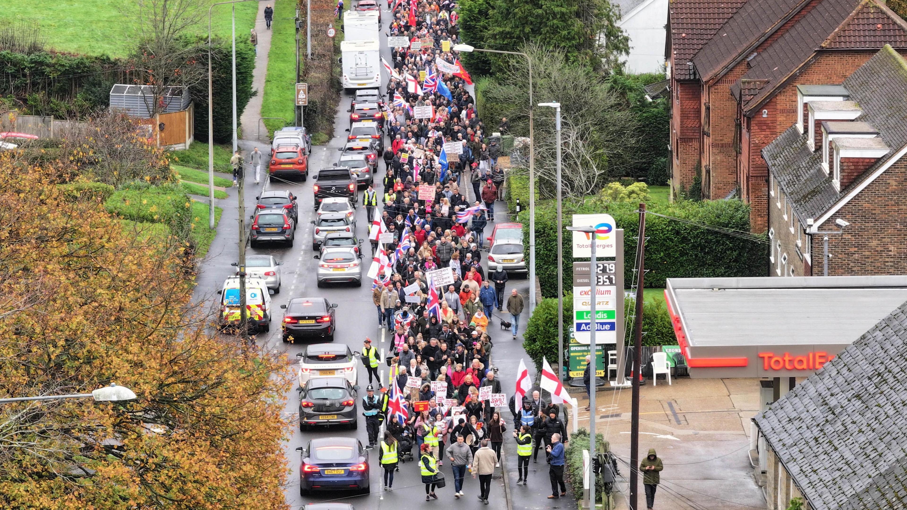 A drone shot of a protest held in Crowborough, East Sussex on 16 November over plans to house asylum seekers at an army site in the town. Hundreds of people can be seen walking on the streets with flags and banners. Cars are driving alongside the walkers. 