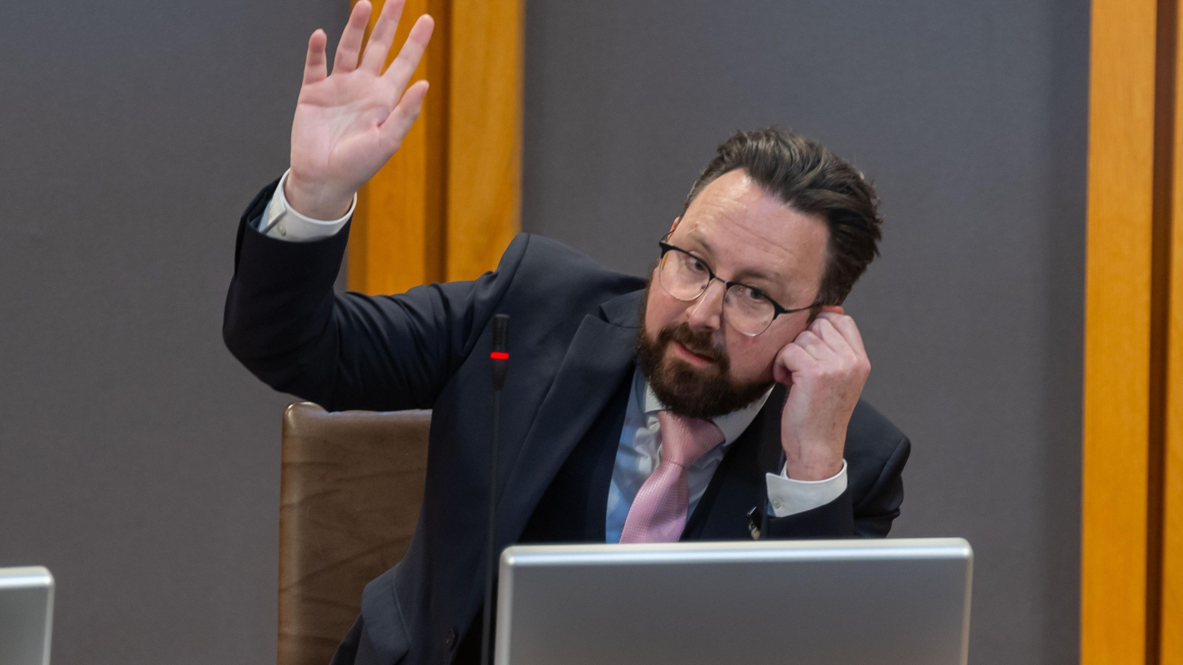 Hefin David sat in a chair with his right arm in the air and his left hand to his ear, wearing a dark suit, pink tie and blue shirt. A computer is in front of him.