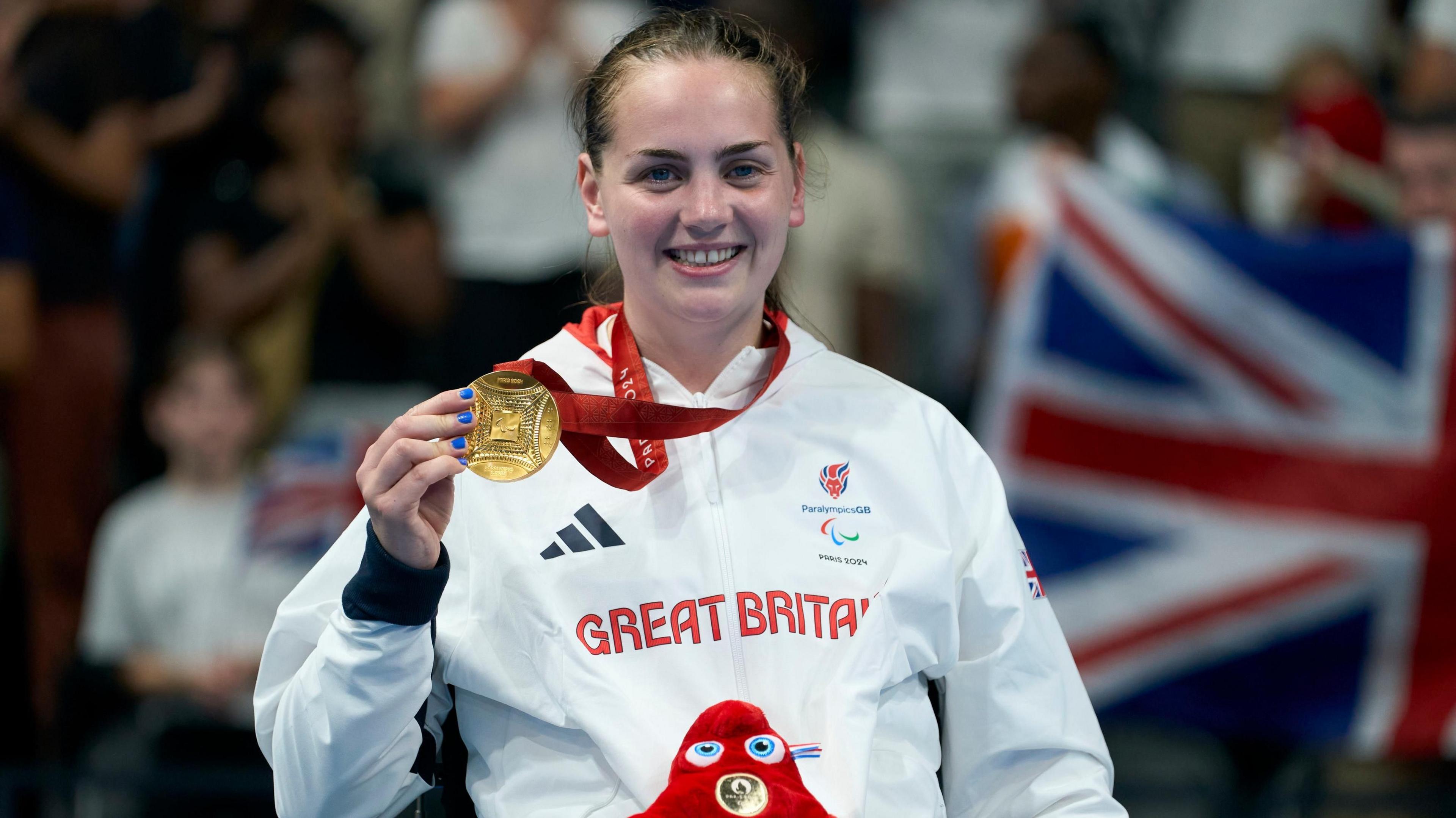 Tully Kearney, a young woman with scraped-back brown hair, smiles as she holds up a gold medal. She is wearing a white Great Britain Paralympics GB jacket.