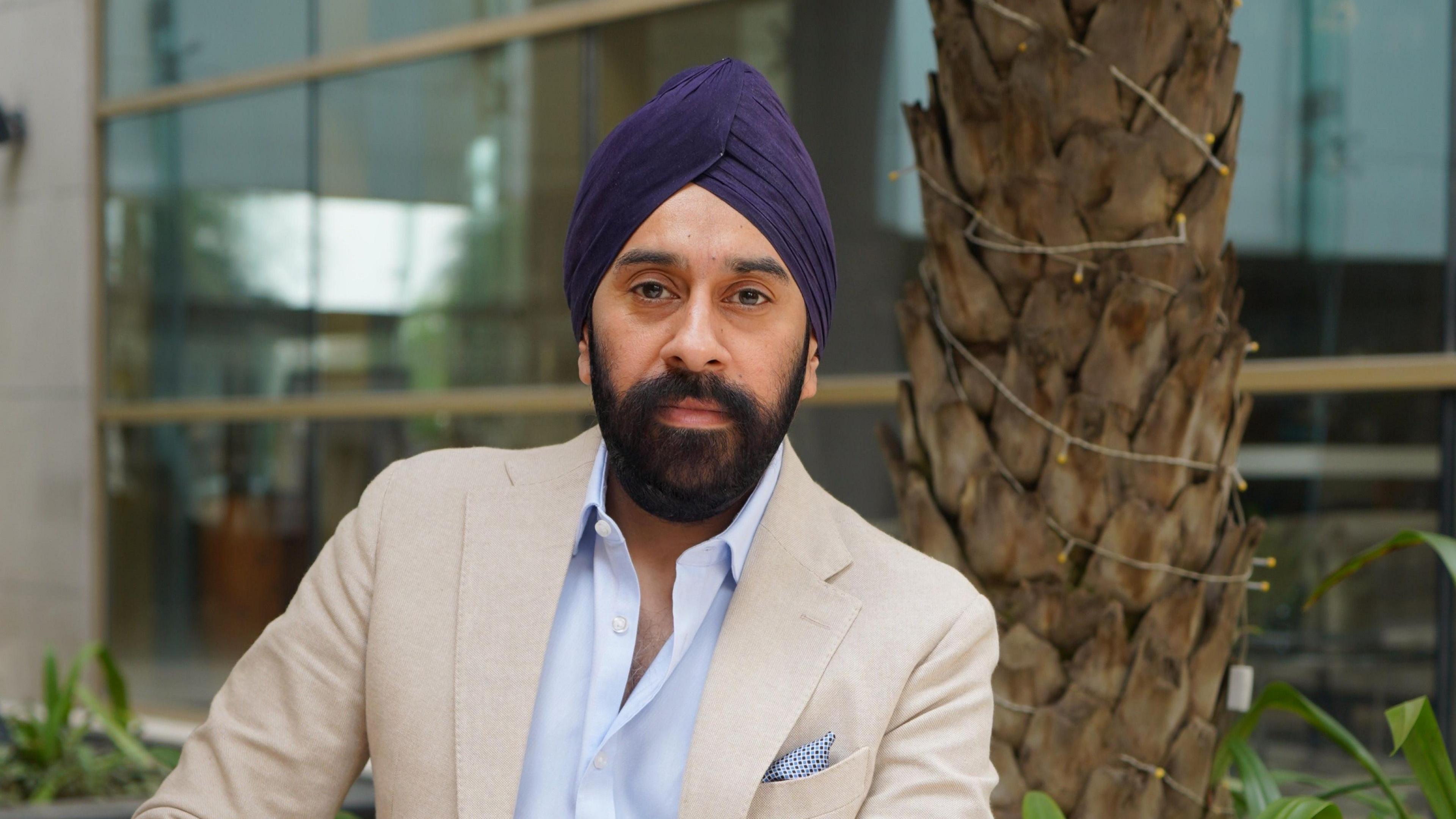 A man wearing a dark purple turban and is sitting down on a chair outside. He is wearing a cream blazer with a baby blue shirt. He is looking directly at the camera.