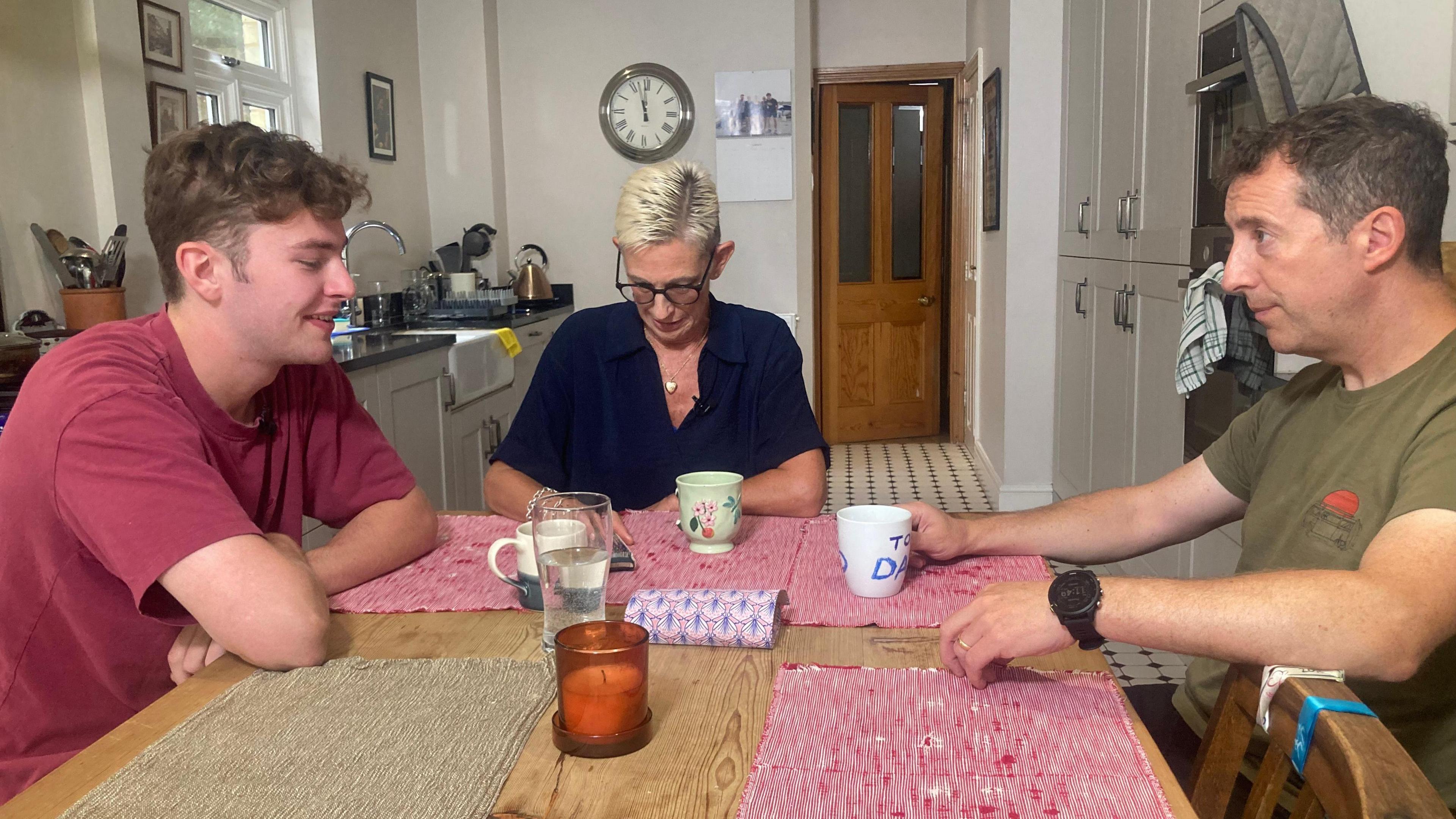 Joe and Juliette Niembro sat at a wooden table in their kitchen with son, Manolo. They are having a conversation looking away from the camera. 
Manolo is wearing a red t-shirt and has his arms crossed resting on the table. Joe is wearing a green t-shirt and is holding a white cup that says Dad in blue letters. Mrs Niembro sits at the end of a table in a navy blouse. She is looking down at her phone and has black square framed glasses on. 