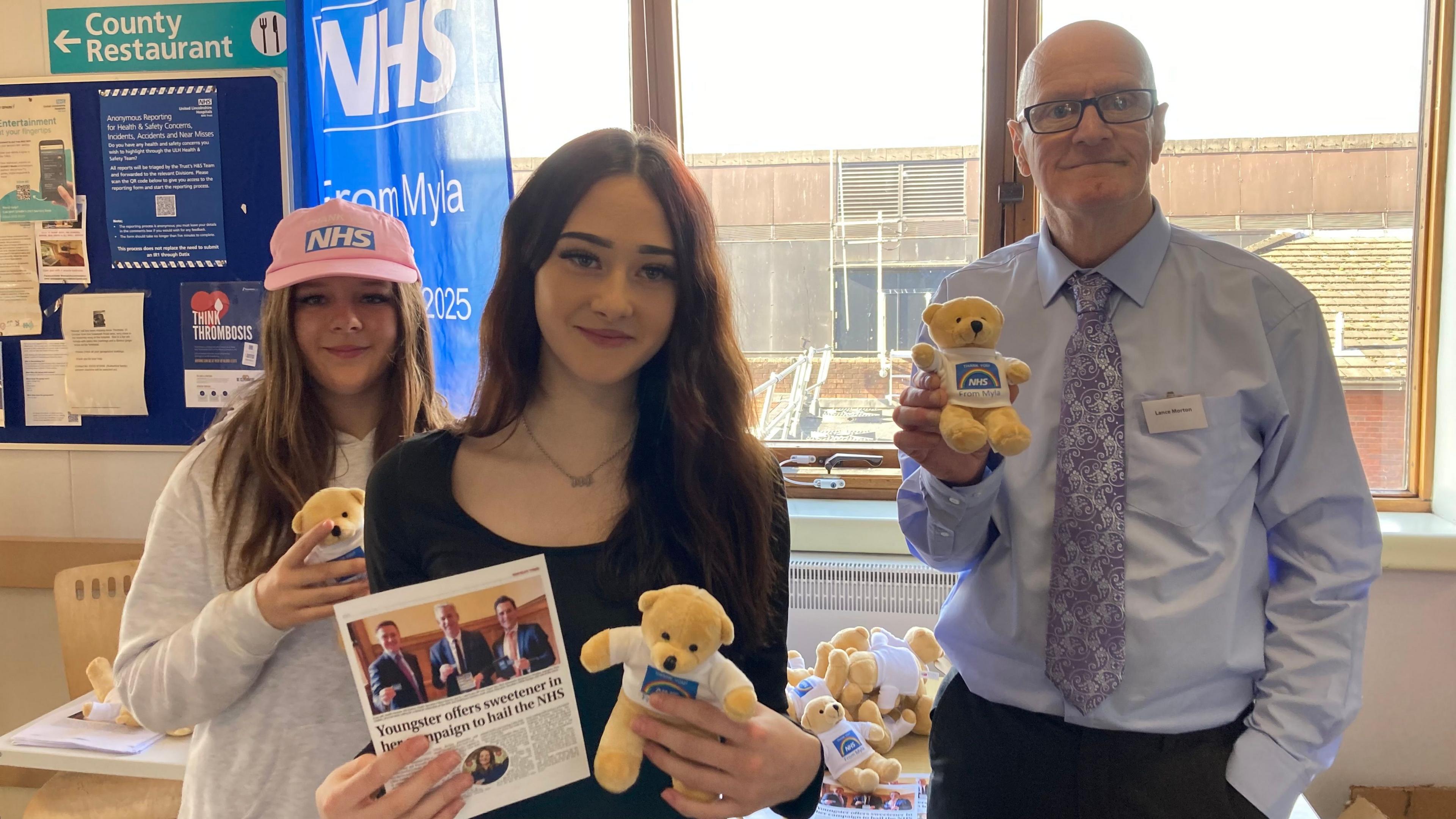 Evie is stood on the left with long brown hair, wearing a grey hoodie and a pink hat with a blue NHS logo. She is holding a yellow bear with a white top on showing the NHS logo. Myla is stood in the middle wearing a black long sleeve dress. She has brown hair and is also holding a yellow bear and a leaflet. Her stepdad is on the right wearing a blue shirt, a purple patterned tie and a name badge. He is bald and is wearing glasses. He is also holding a bear. They are all standing in a hospital in front of a window.