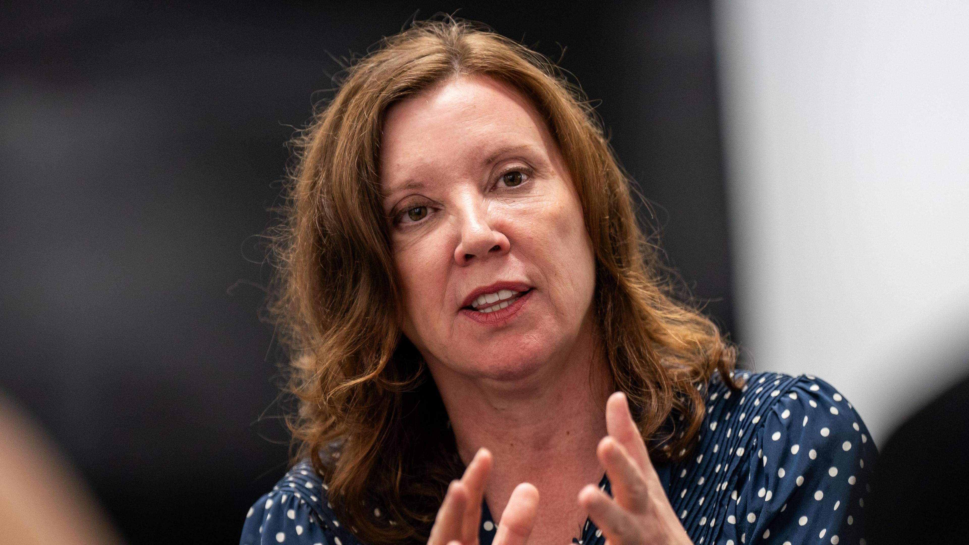 Woman with brown hair and blue top with white spots centre frame, she is looking up and to the right with her hands held up to her shoulders.