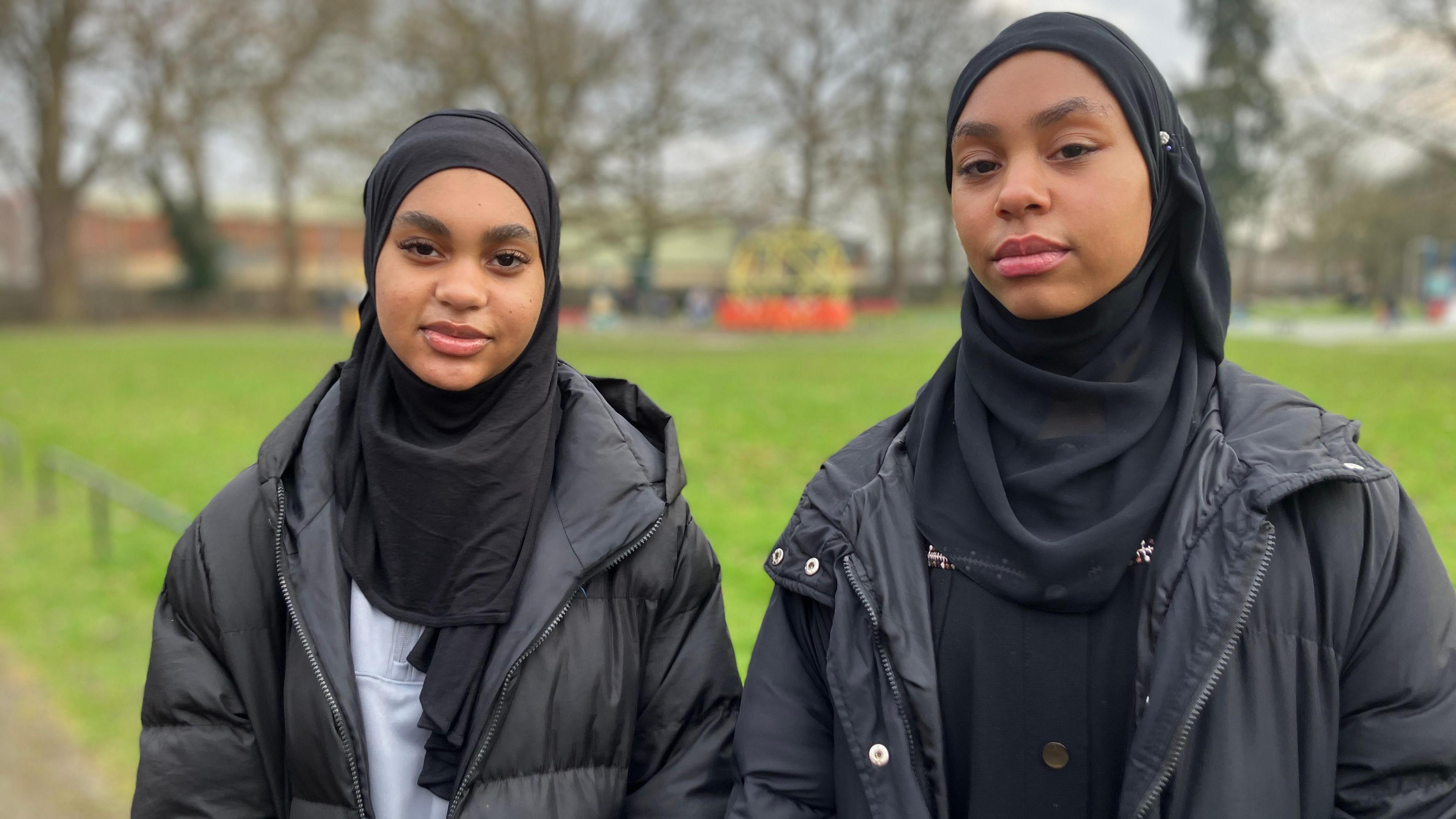 Innat and Hanan stand in a park. The background is blurred, but there is grass, a small children's climbing frame play area and trees lining the edge of the park. The girls both look at the camera. Innat wears a black hijab, with a black hooded puffer coat and a pale grey zipped sweatshirt underneath. Hanan also wears a black hijab, with a hooded black puffer jacket and a black buttoned blouse underneath.