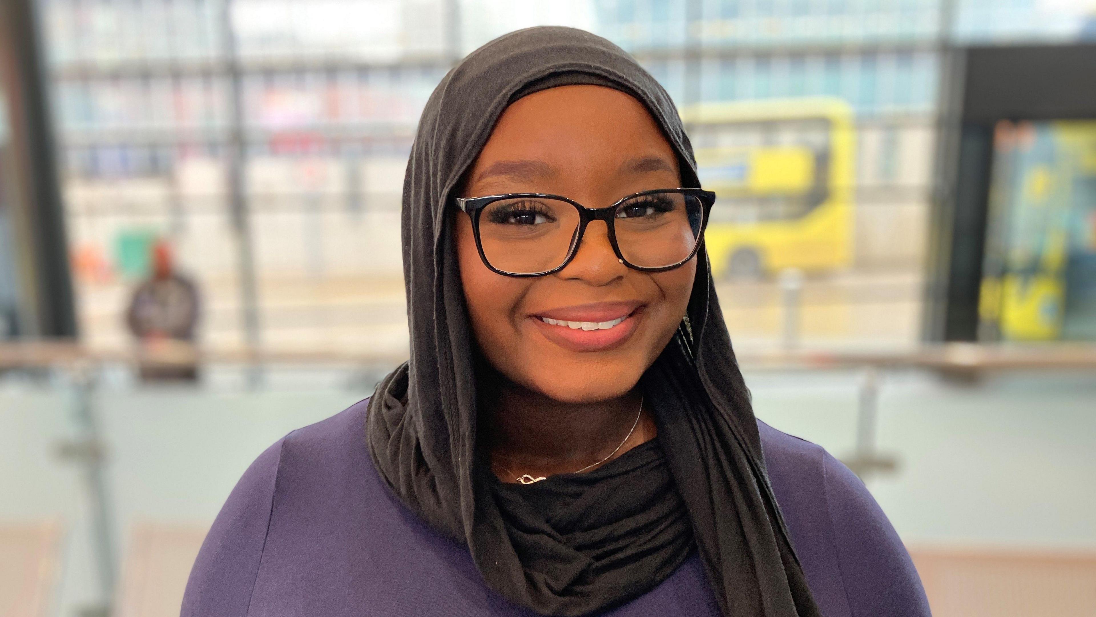 A smiling, young woman wearing black glasses and a hijab.