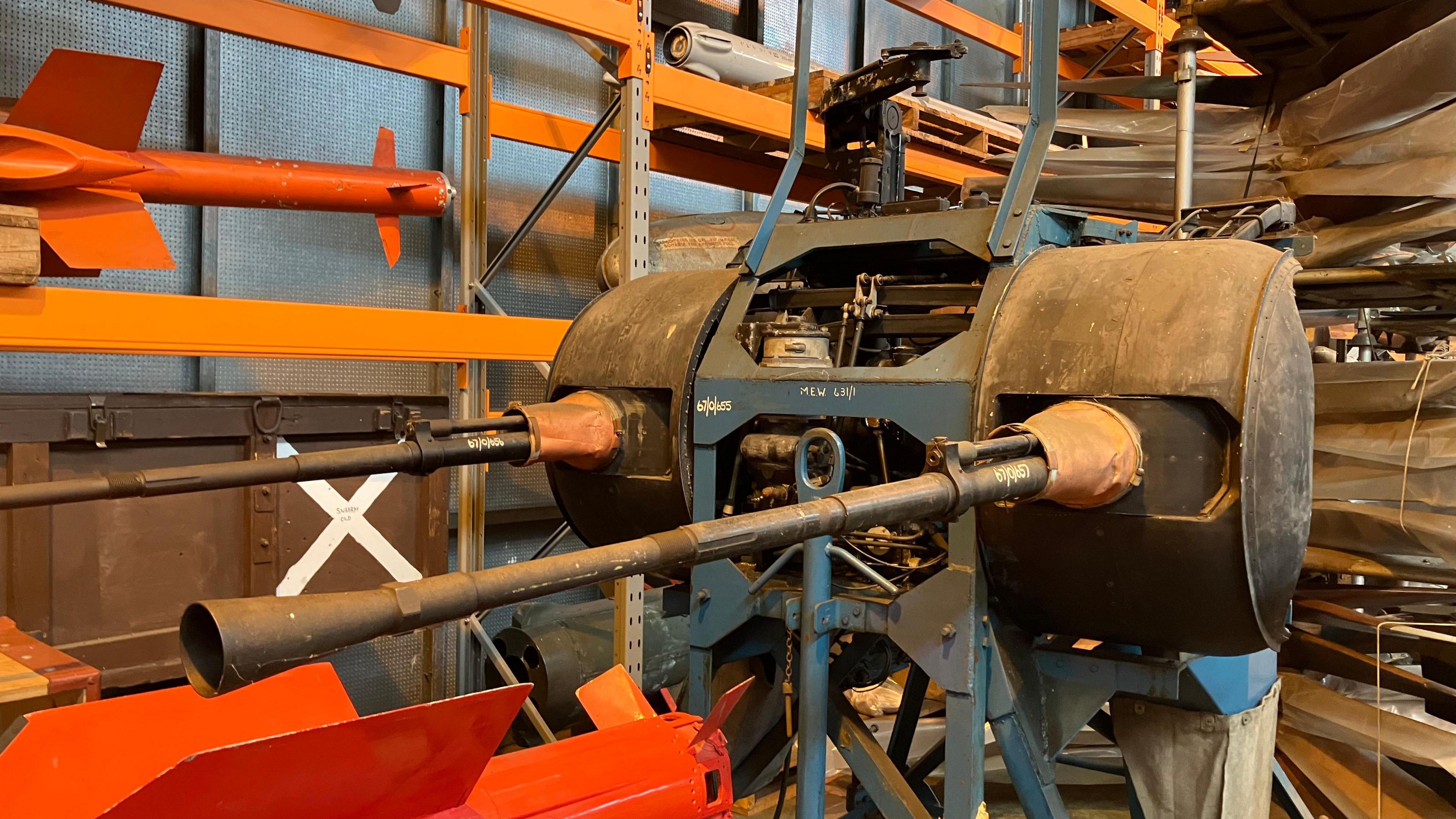 An large old gun that used to be on an RAF aircraft. It has been stored in a large warehouse, near some shelving units.