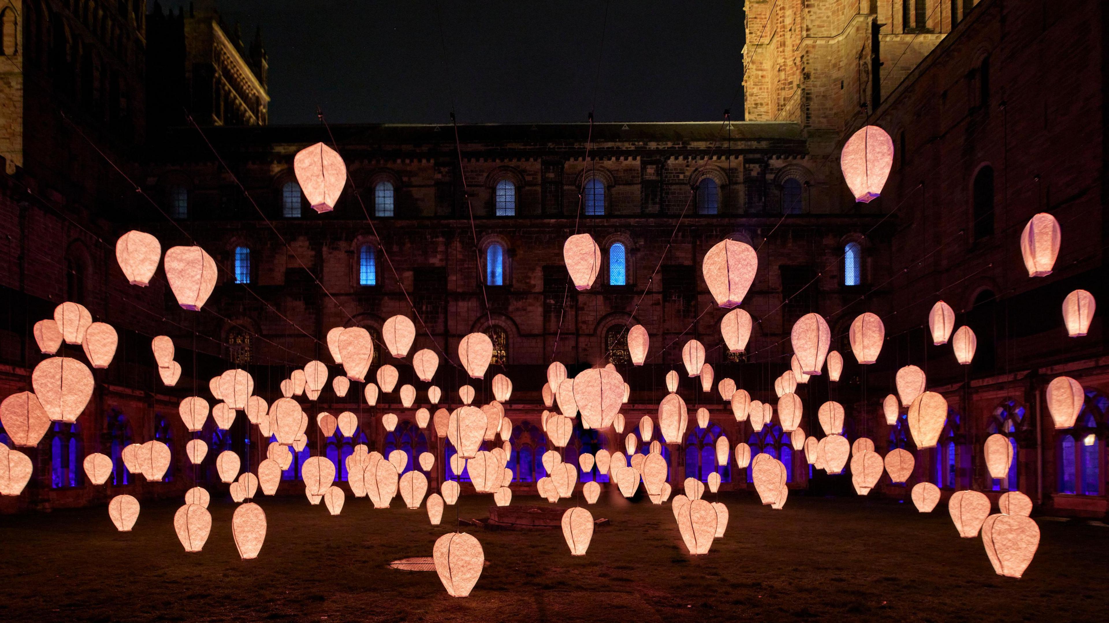 Dozens of paper lanters shining in a peach colour are floating in Durham Cathedral's Cloister in artwork Solace.