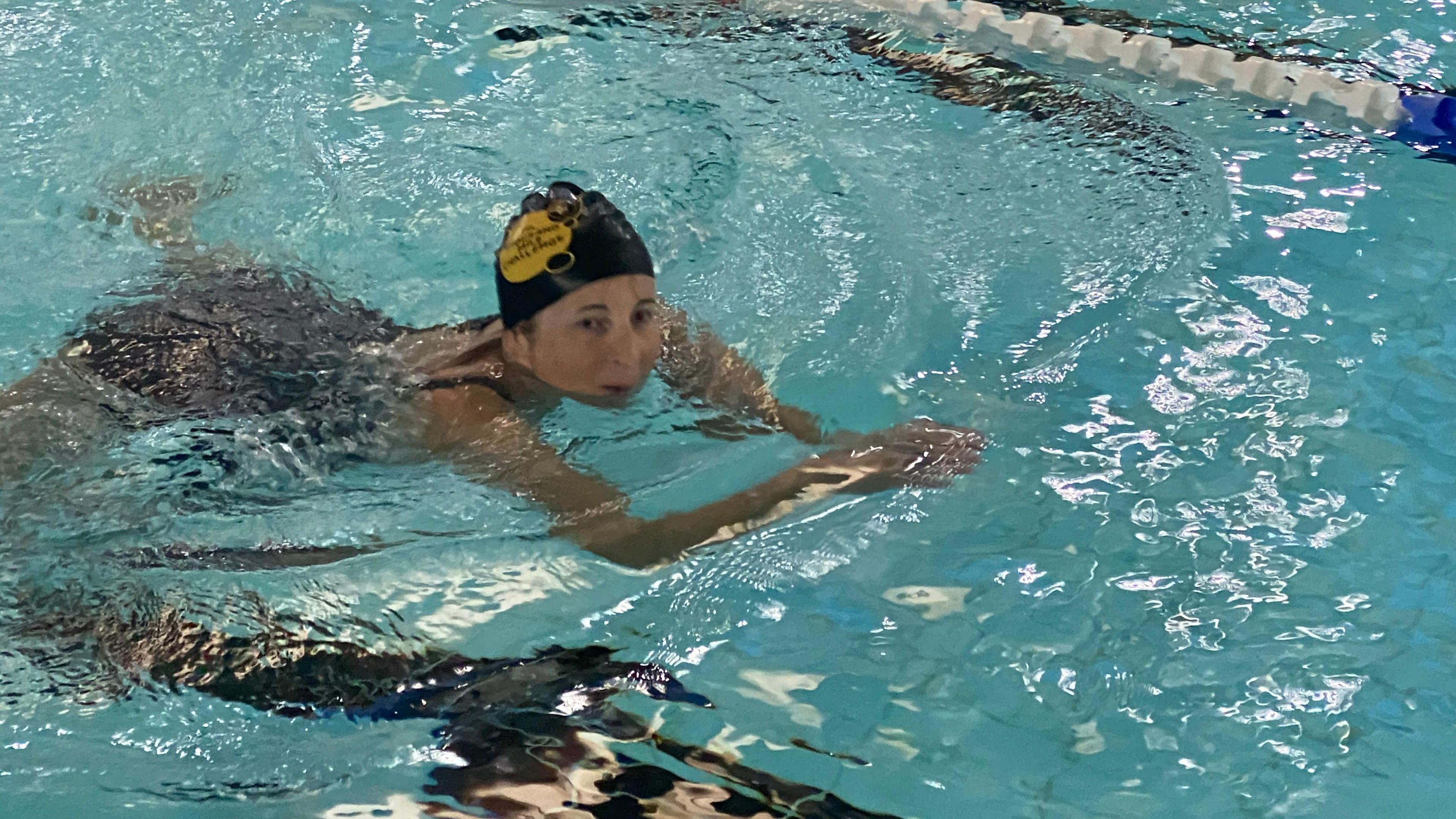 Kate is wearing the Thousand Mile Challenge while doing the breaststroke in the pool and she is wearing a dark blue swimming costume. She's looking out of the water to her left.