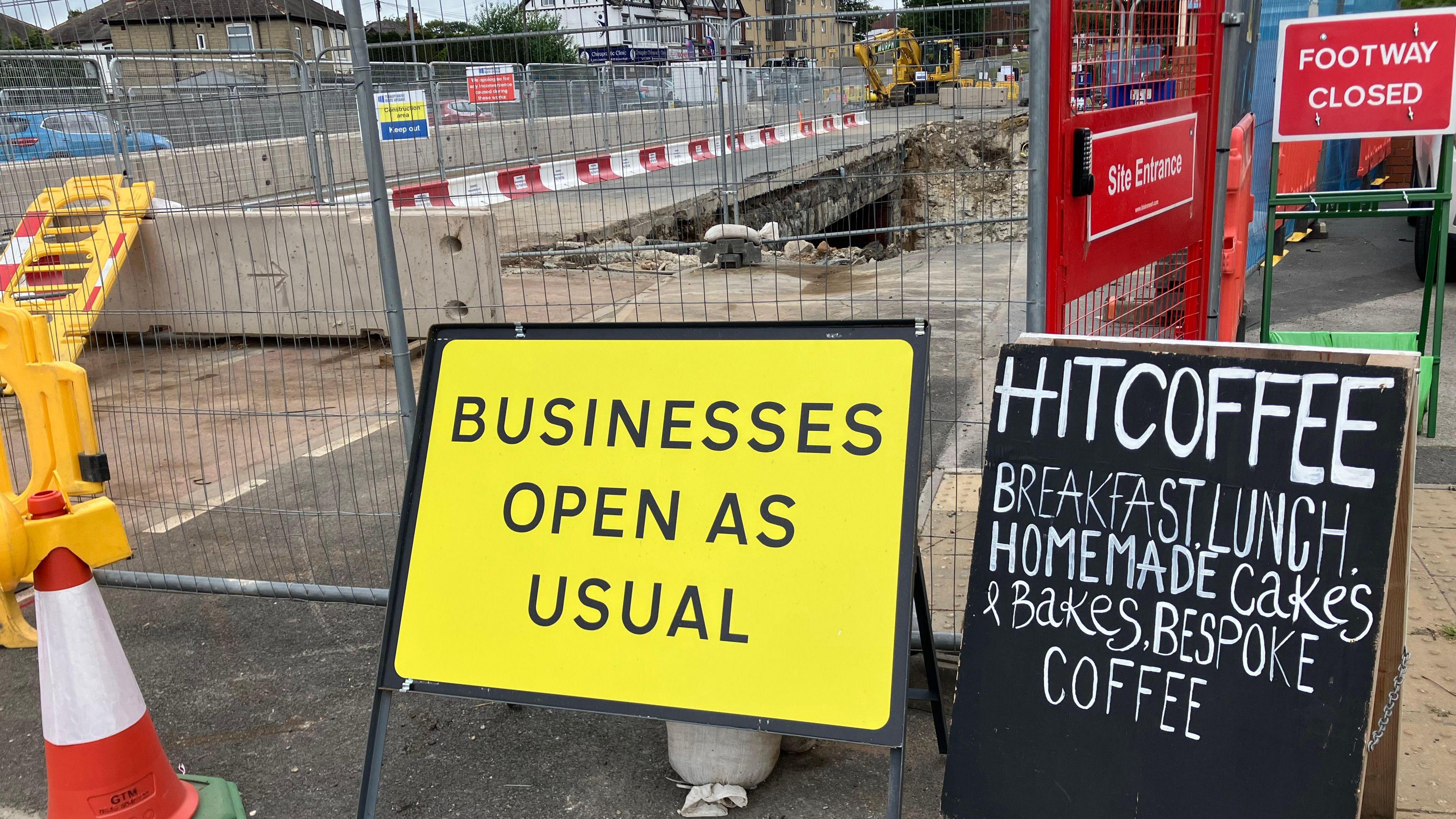 Two signs are displayed in front of major roadworks. One reads "businesses open as usual" on a yellow background, the other reads "HIT COFFEE, breakfast, lunch, homemade cakes & bakes, bespoke coffee".