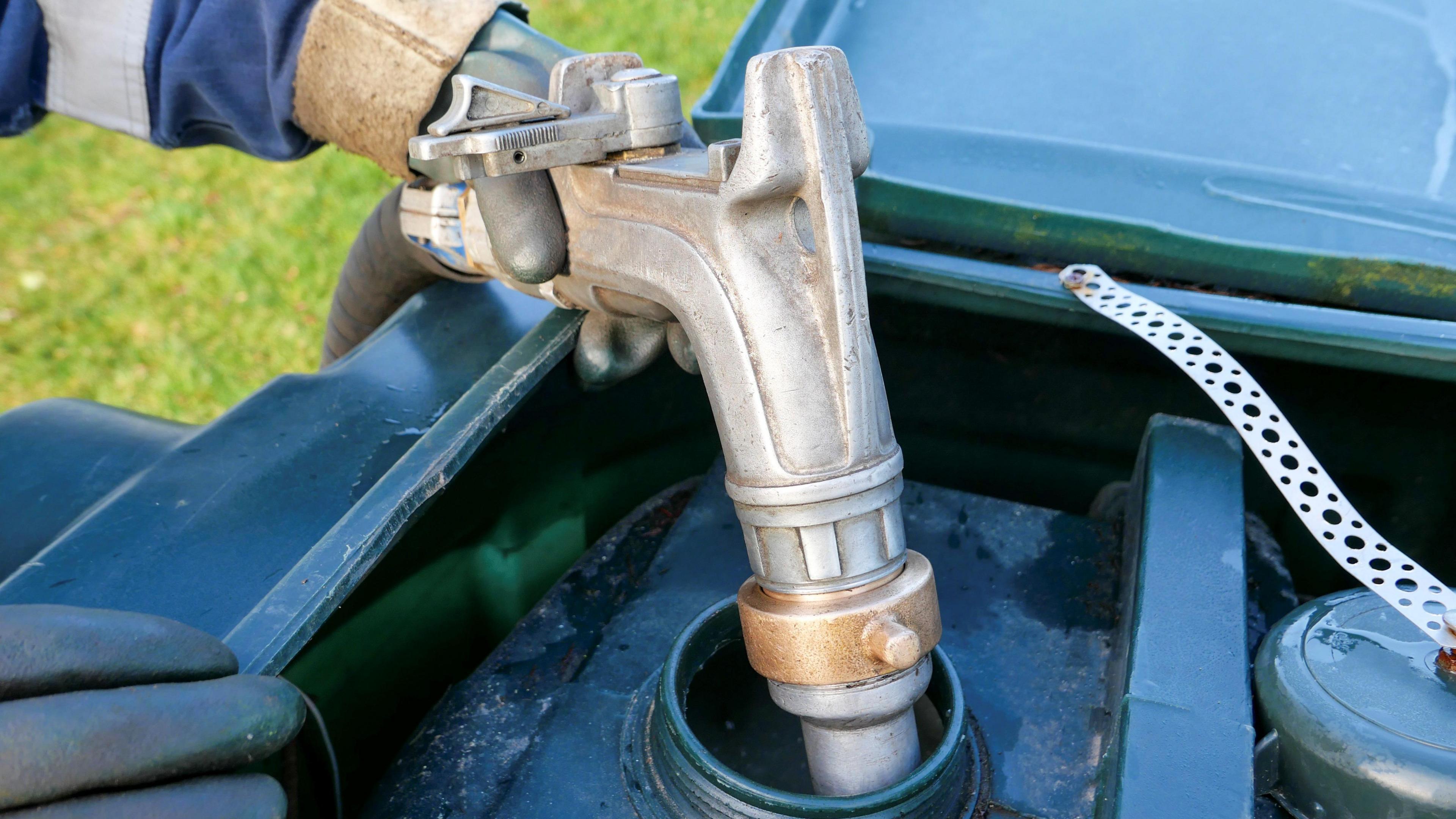 An oil delivery driver filling up a residential oil tank.  The mains gas is not connected. The man is wearing protective overalls and gloves as he carefully fills the green plastic tank with home heating oil, in preparation for the winter months. He is squeezing a metal nozzle attached to a long rubber hose that runs from his oil delivery lorry parked nearby to maintain control of the amount being delivered.