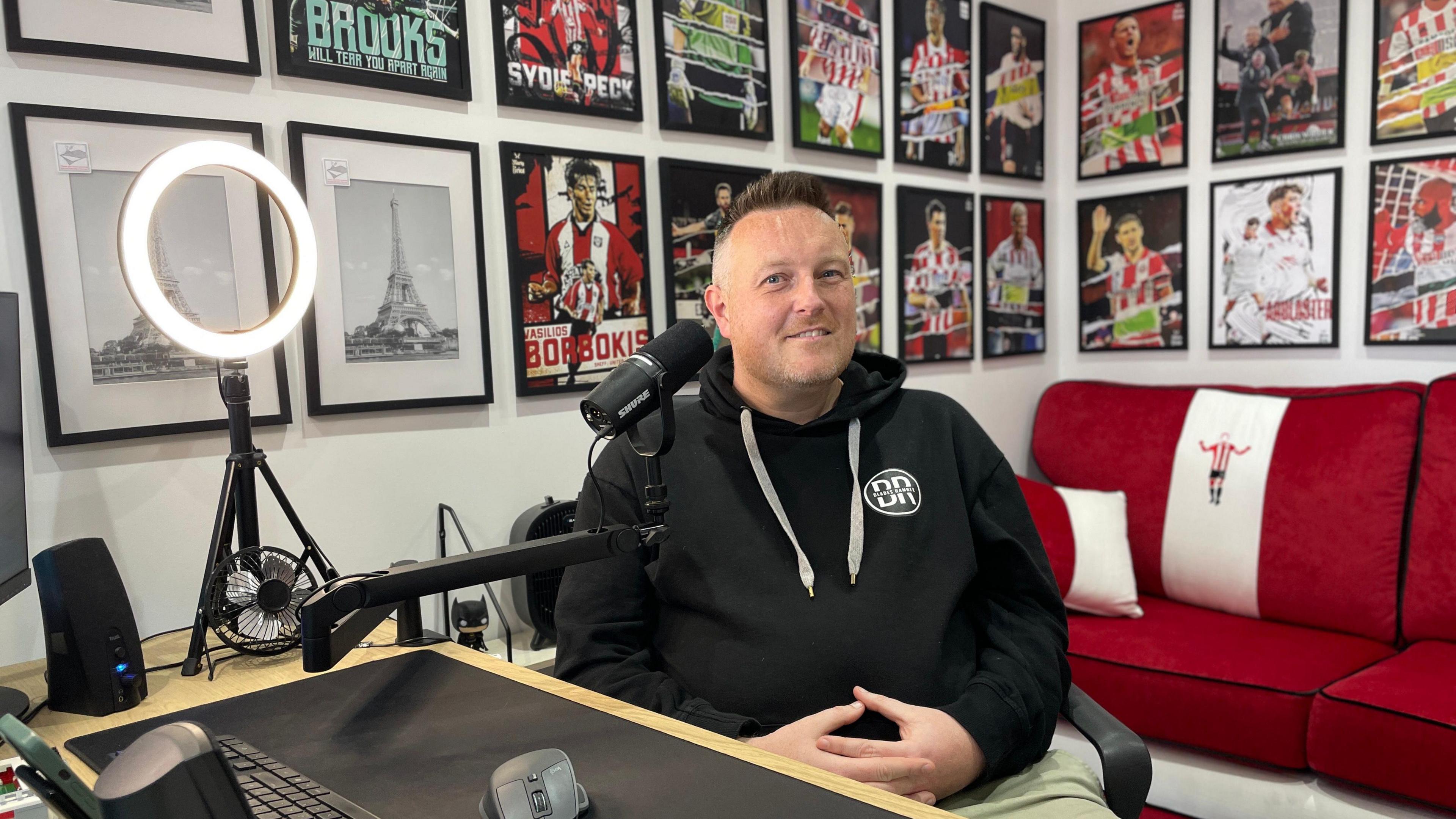 A man with short hair and wearing a dark hoodie sat in a recording studio. A mic is close to his face. Behind him is a red sofa and posters of footballers on the walls, as well as some pictures of the Eiffel Tower.
