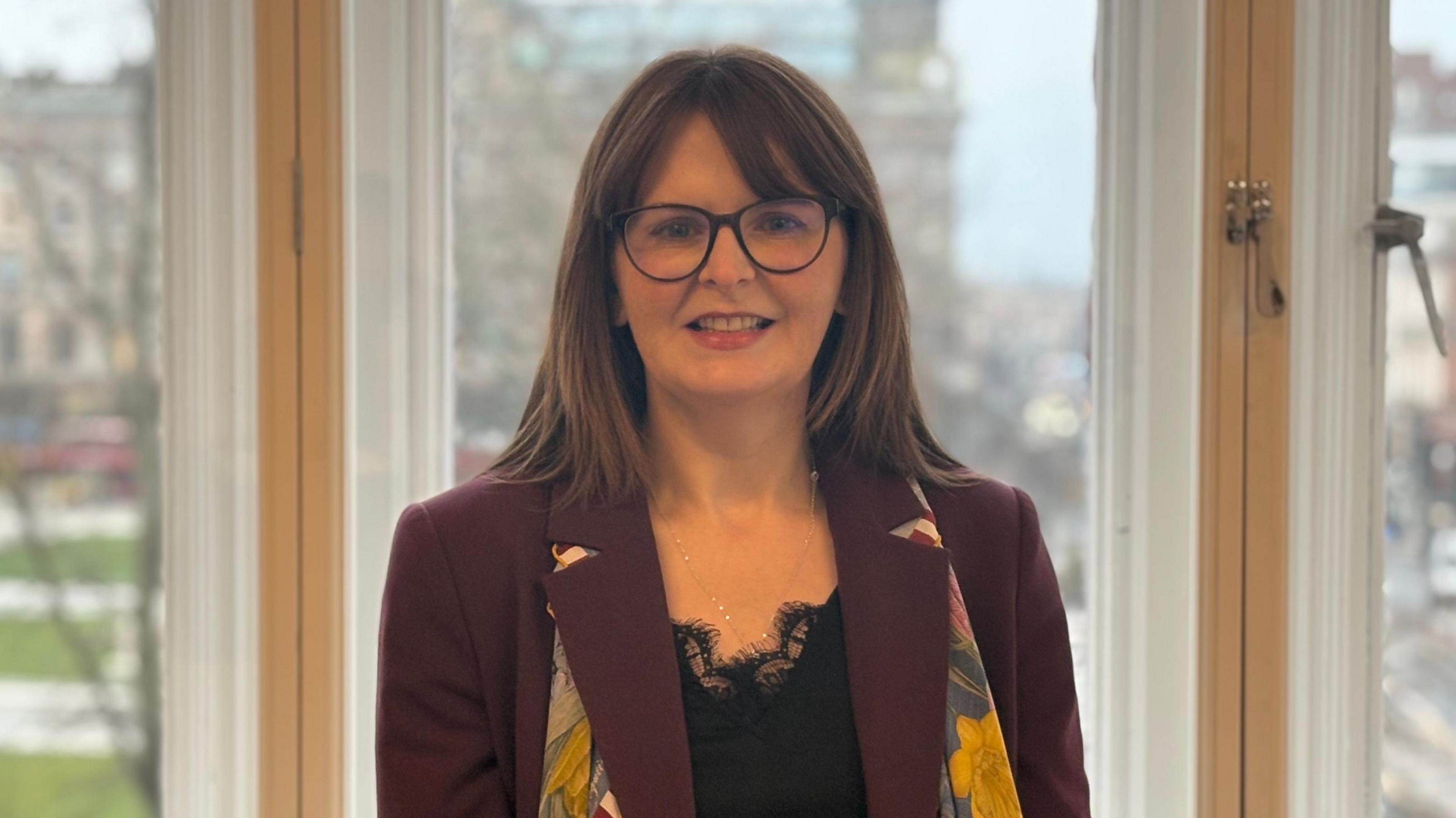 Caoimhe Archibald is standing in front of large windows and is smiling at the camera. She has shoulder-length hair and is wearing a pair of large-framed glasses. She is also wearing a burgundy coat, a black top and a floral patterned scarf.