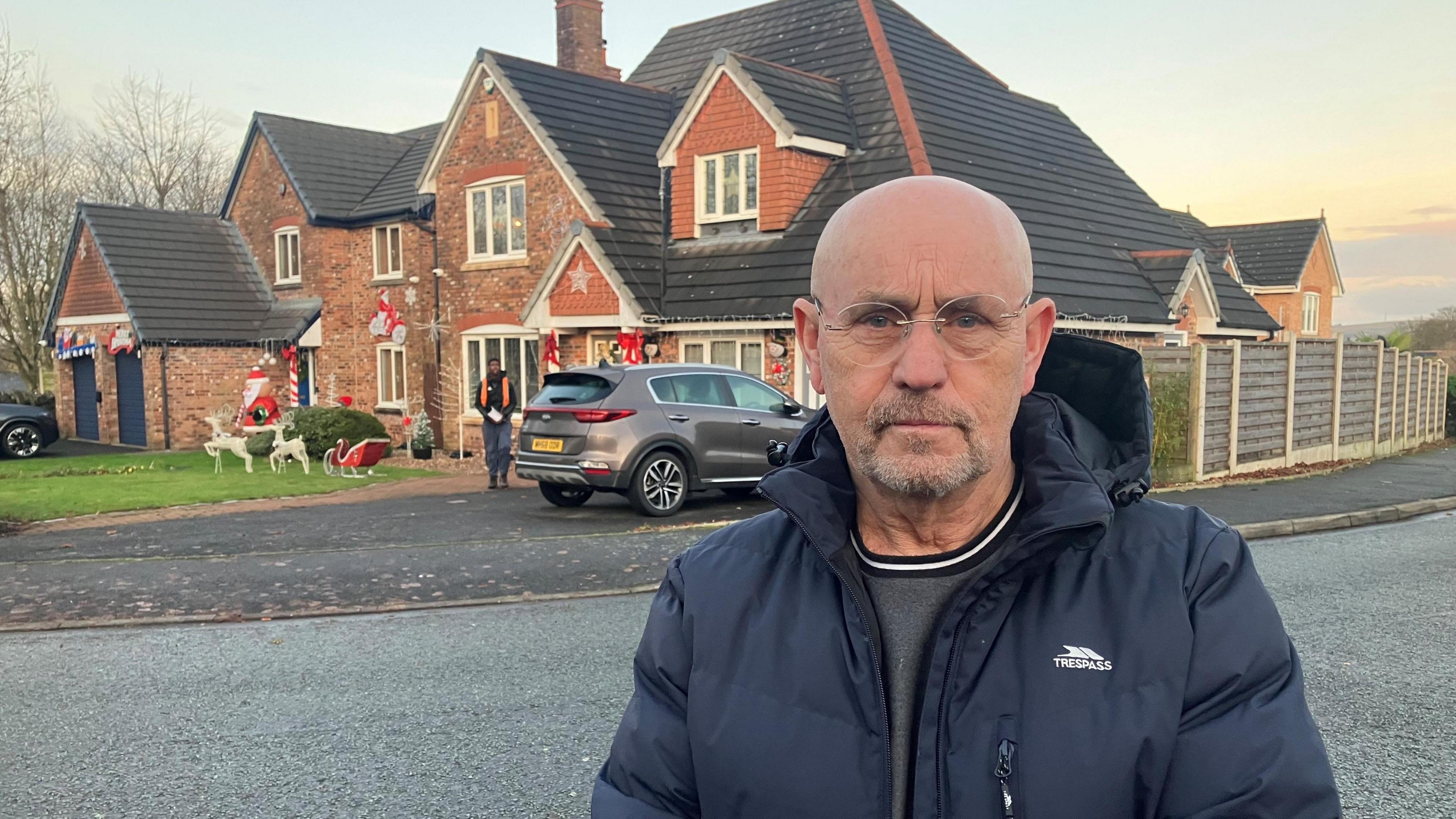 Peter Marr is wearing glasses, a grey T-shirt and a blue padded jacket. He is standing in front of a decorated home in the daytime.