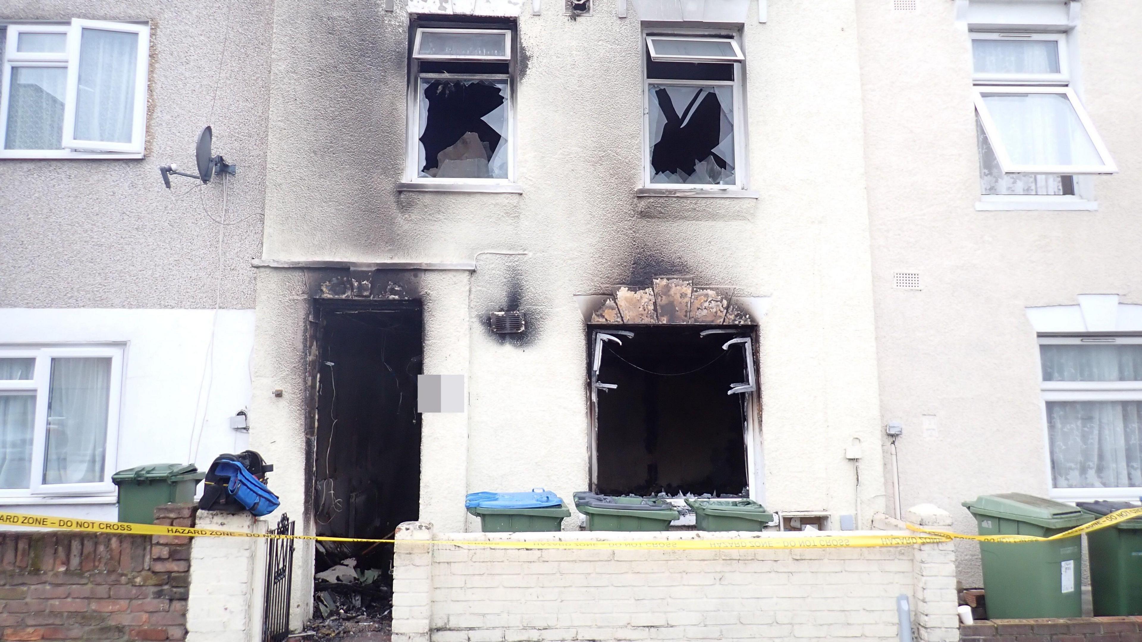 A two-storey terraced house with blackened and broken windows and severe damage to the front of the property. There is a 'do not cross' tape in front of the property. The houses on either side appear undamaged. 