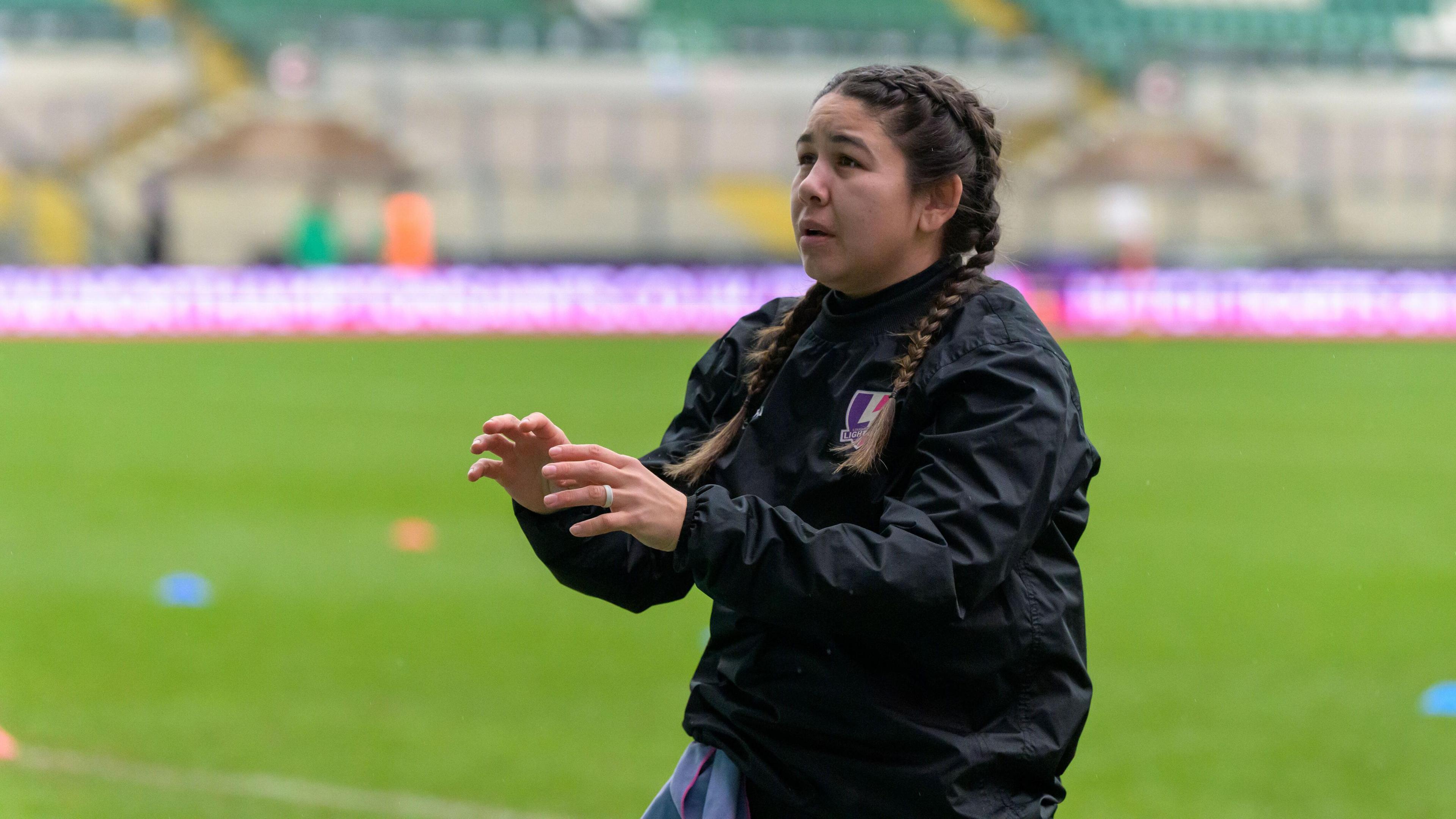 A woman with two plaits in her hair is facing the left with her hands up ready to catch a ball. She is wearing a black waterproof rugby coat. She also has a silver ring on her left index finger.