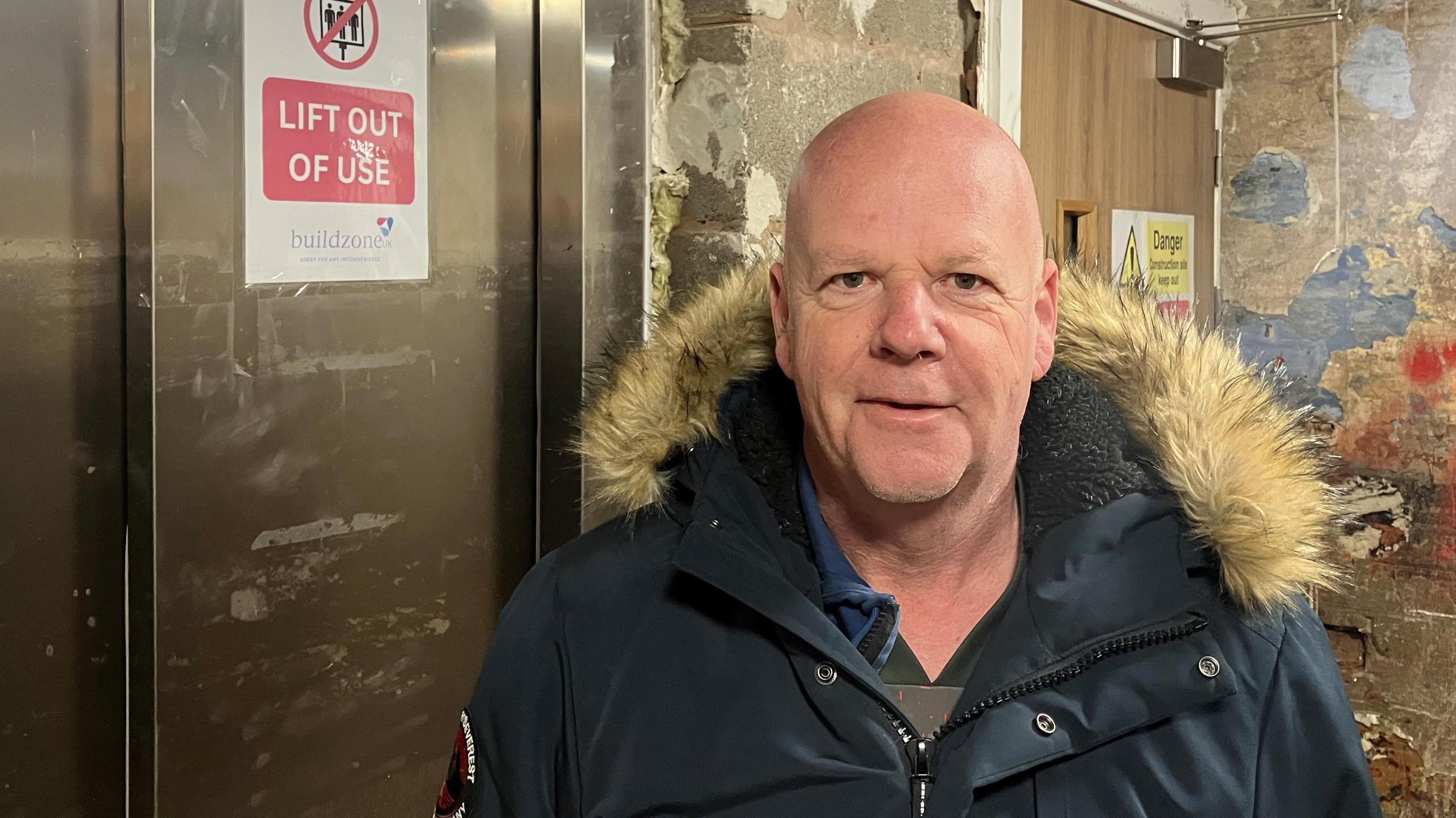 Meadow Mill resident David Howarth, who is bald and wearing a fur-lined parka coat, stands next to an out-of-action lift in the basement of the apartment complex. 