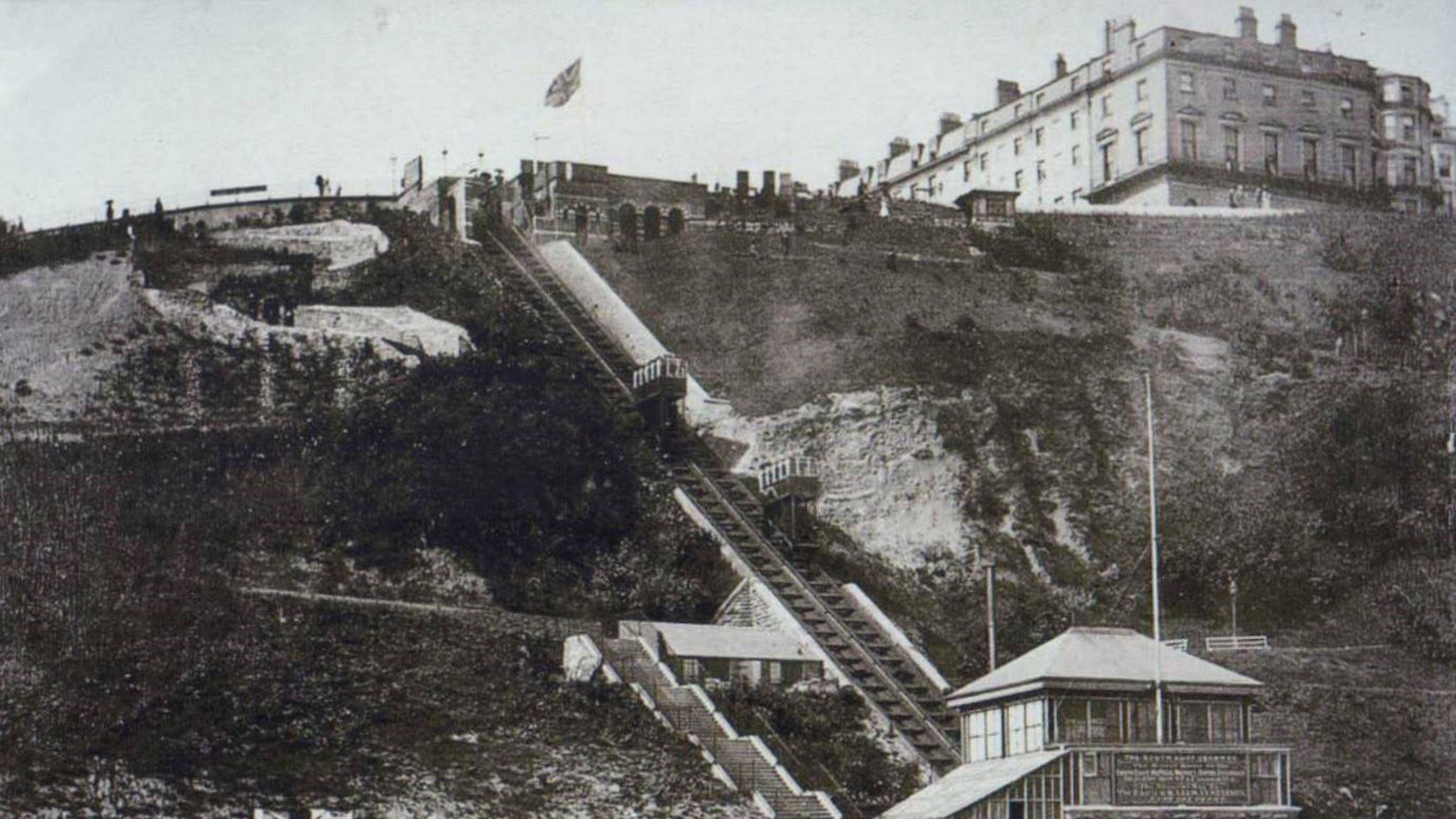 An old black and white image of Scarborough's funicular railway. Railway tracks run vertically up a steep cliff with two buildings at the bottom and top of the track. To the left at the top of the hill, a large Victorian-era hotel can be seen.