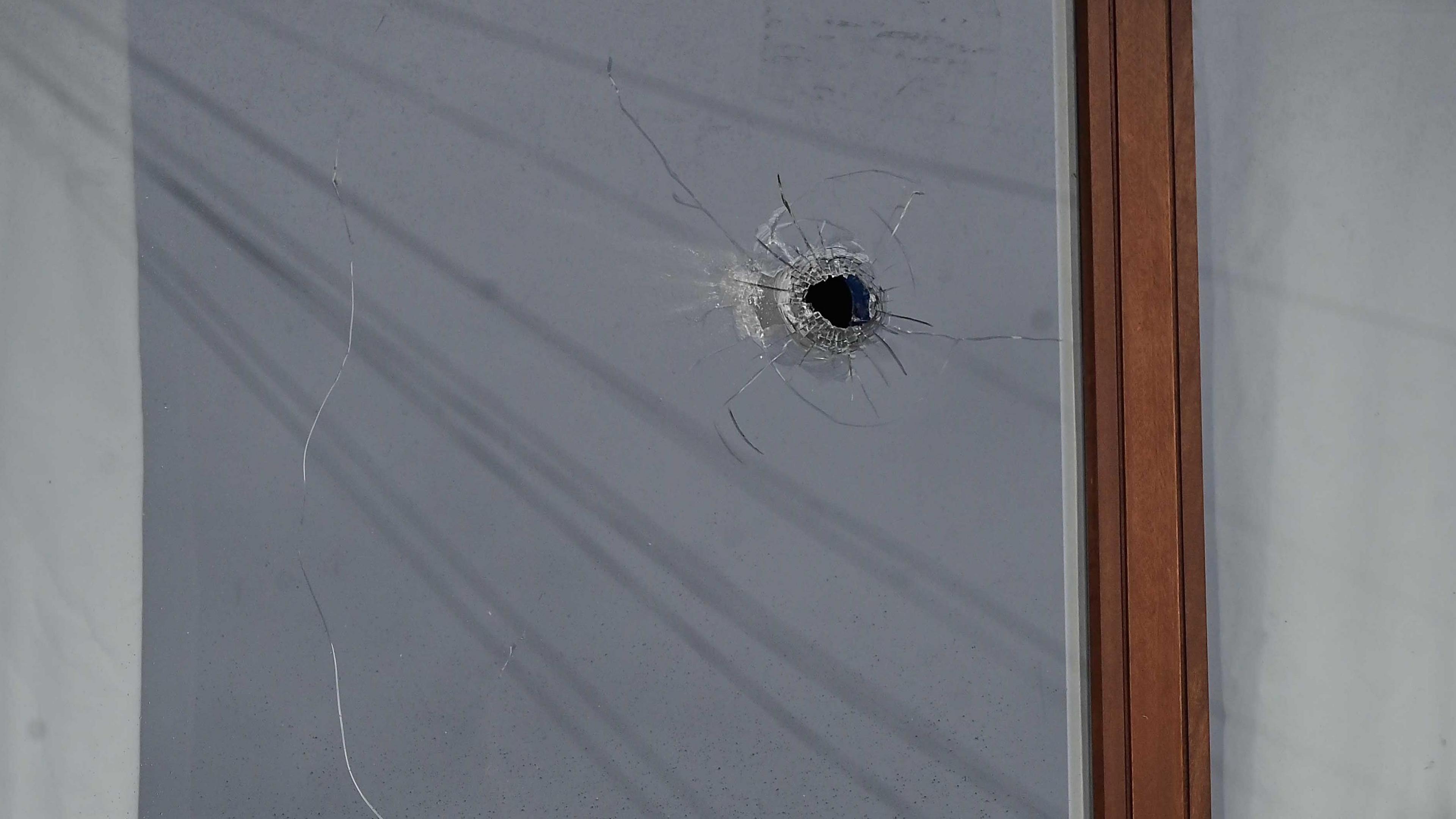 The photo is a close-up of a bullet hole in the window of a house. The window frame is brown wood. 