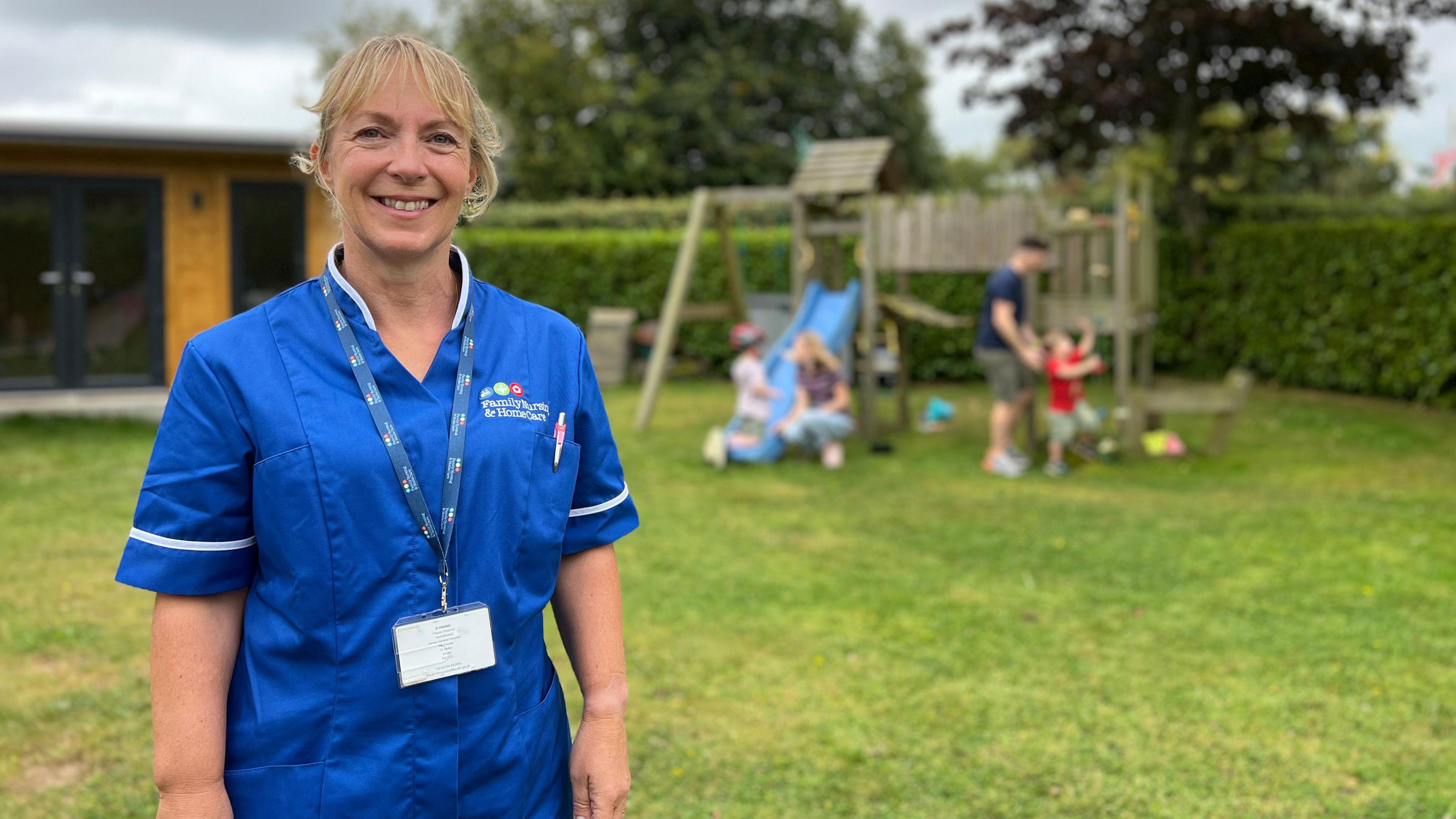 Lynsey is in a blue uniform with a name badge and lanyard stands in the foreground. Behind them, children play on a slide and swing set in a grassy outdoor playground with trees and a building in the background.