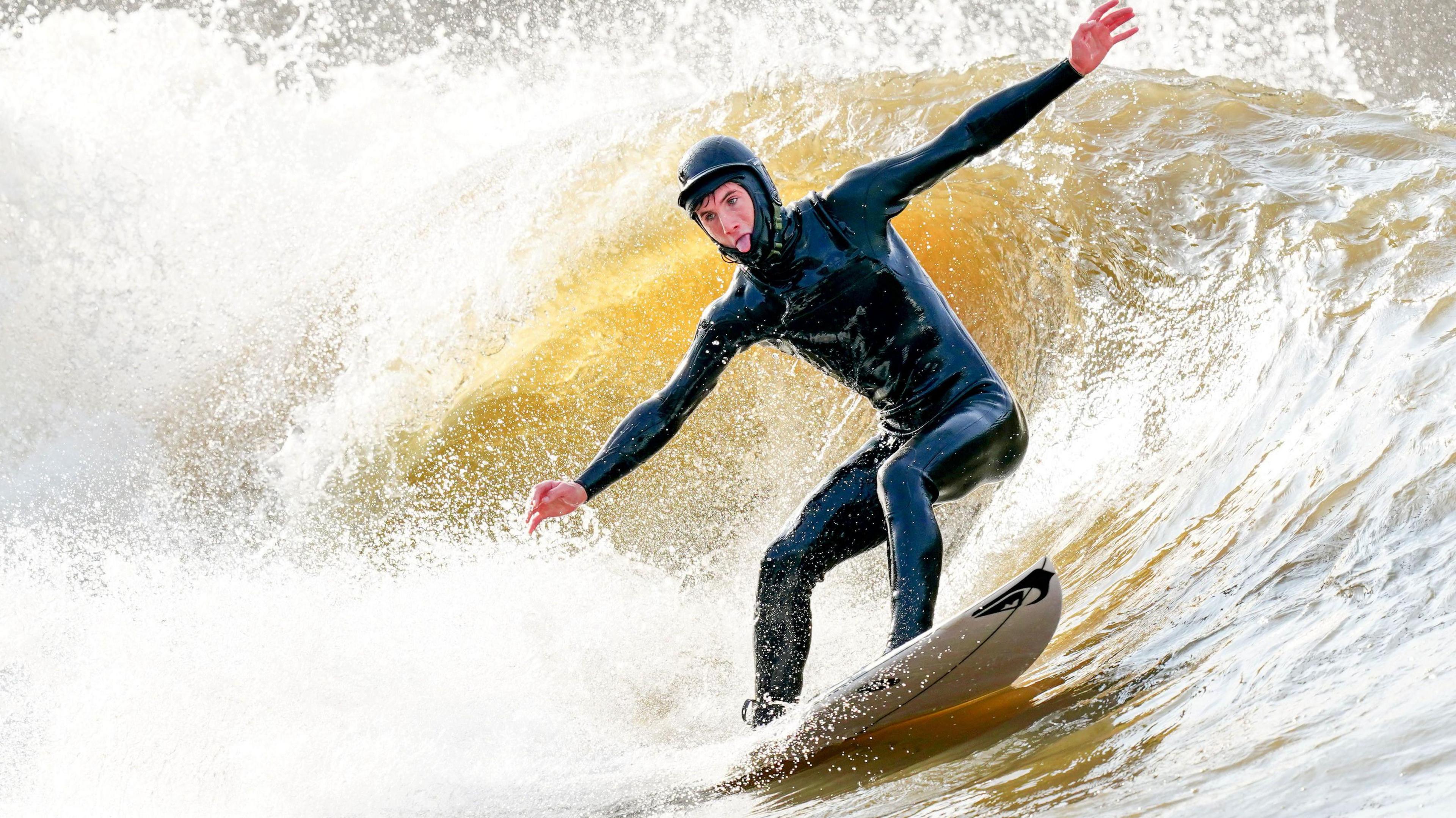 Someone wearing a black wetsuit and black helmet surfs a wave with his arms outstretched