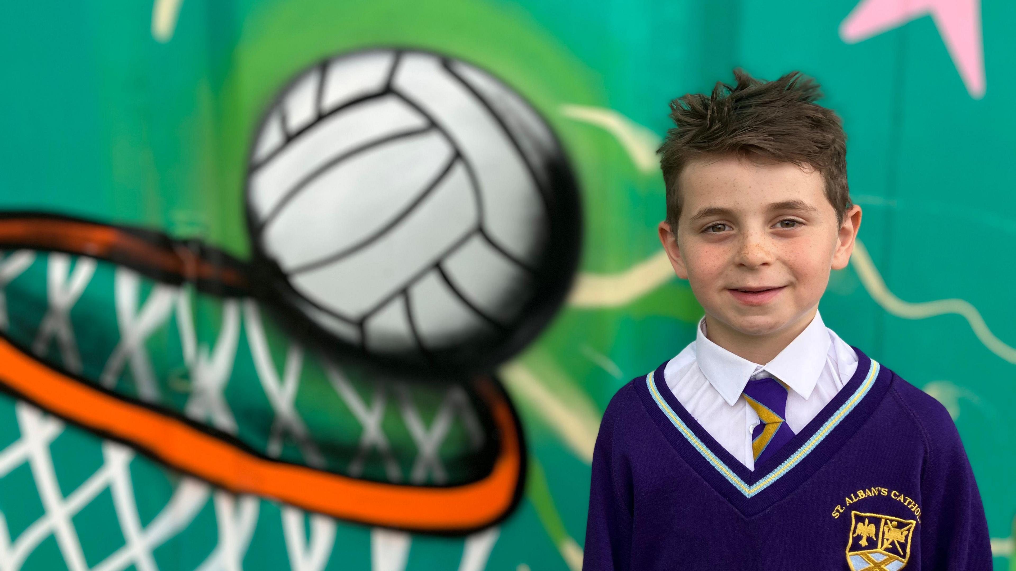 A seven-year-old boy called Bertie, who has brown hair and wears a purple school uniform and tie, smiles at the camera. He stands in front of a mural of a basketball falling into a hoop.