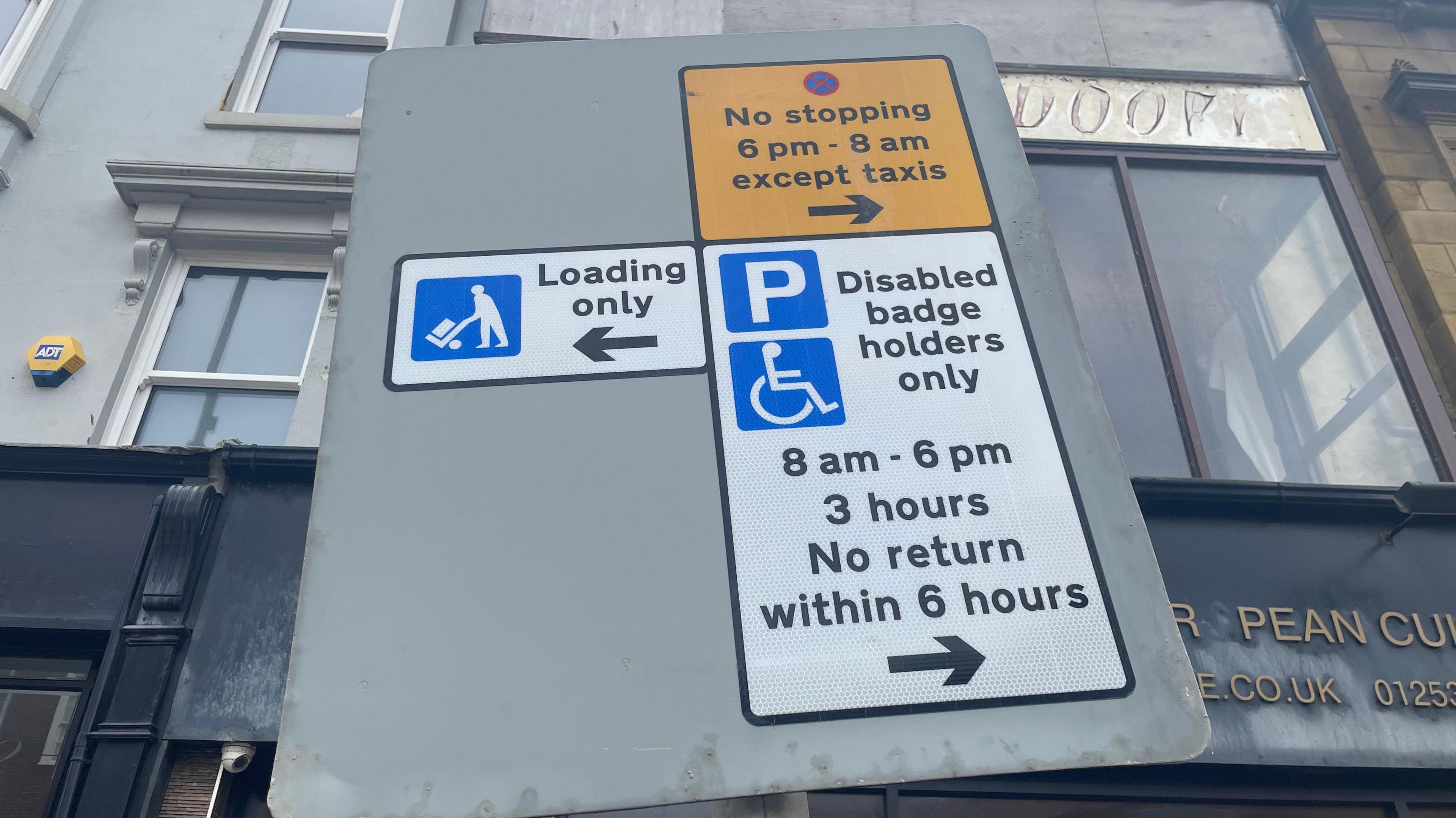 A grey street sign showing the disabled parking spaces and also the sign for when it becomes a taxi rank.