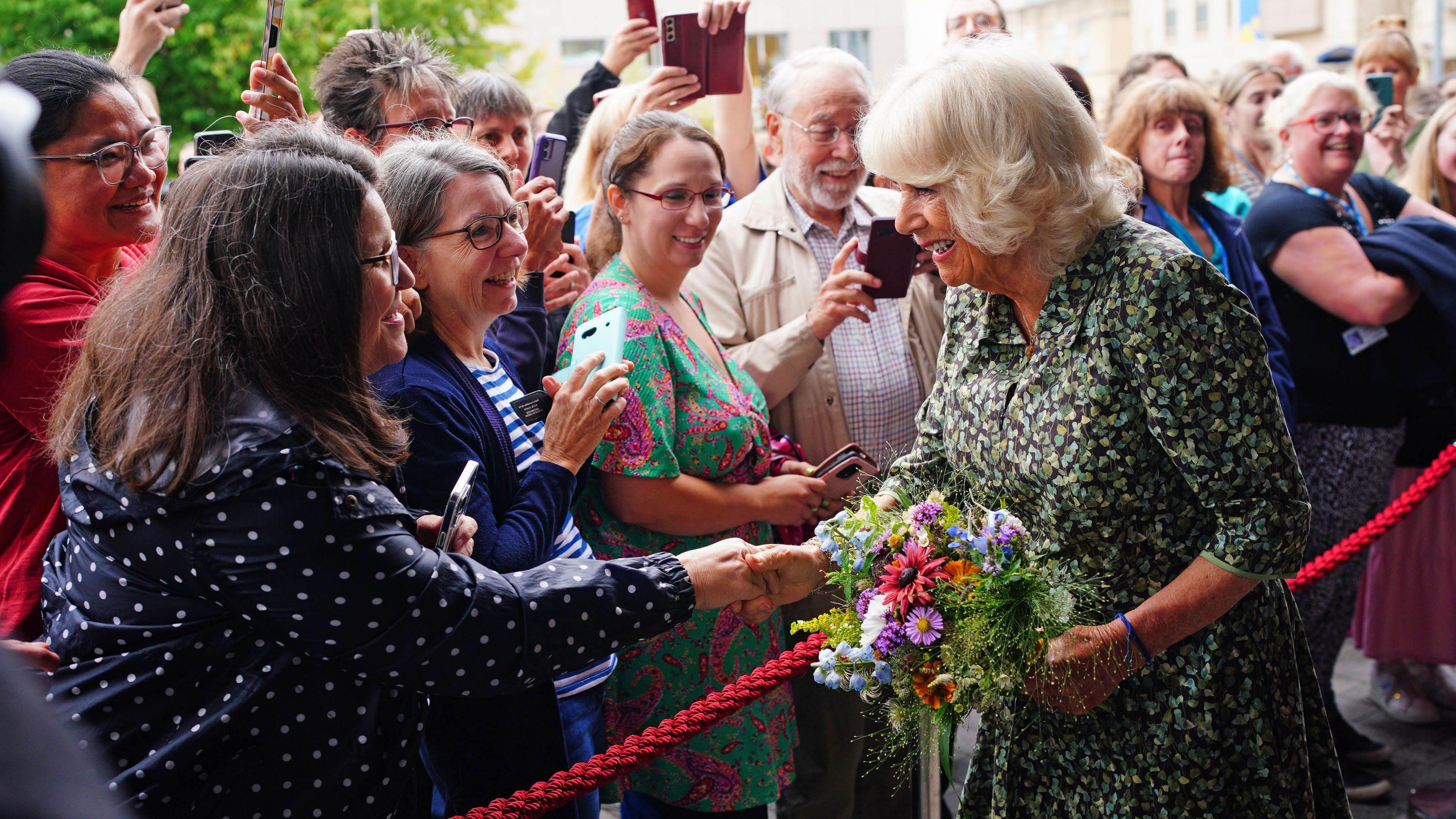 The Queen holding flowers and shaking hands with the public who are standing behind a red rope