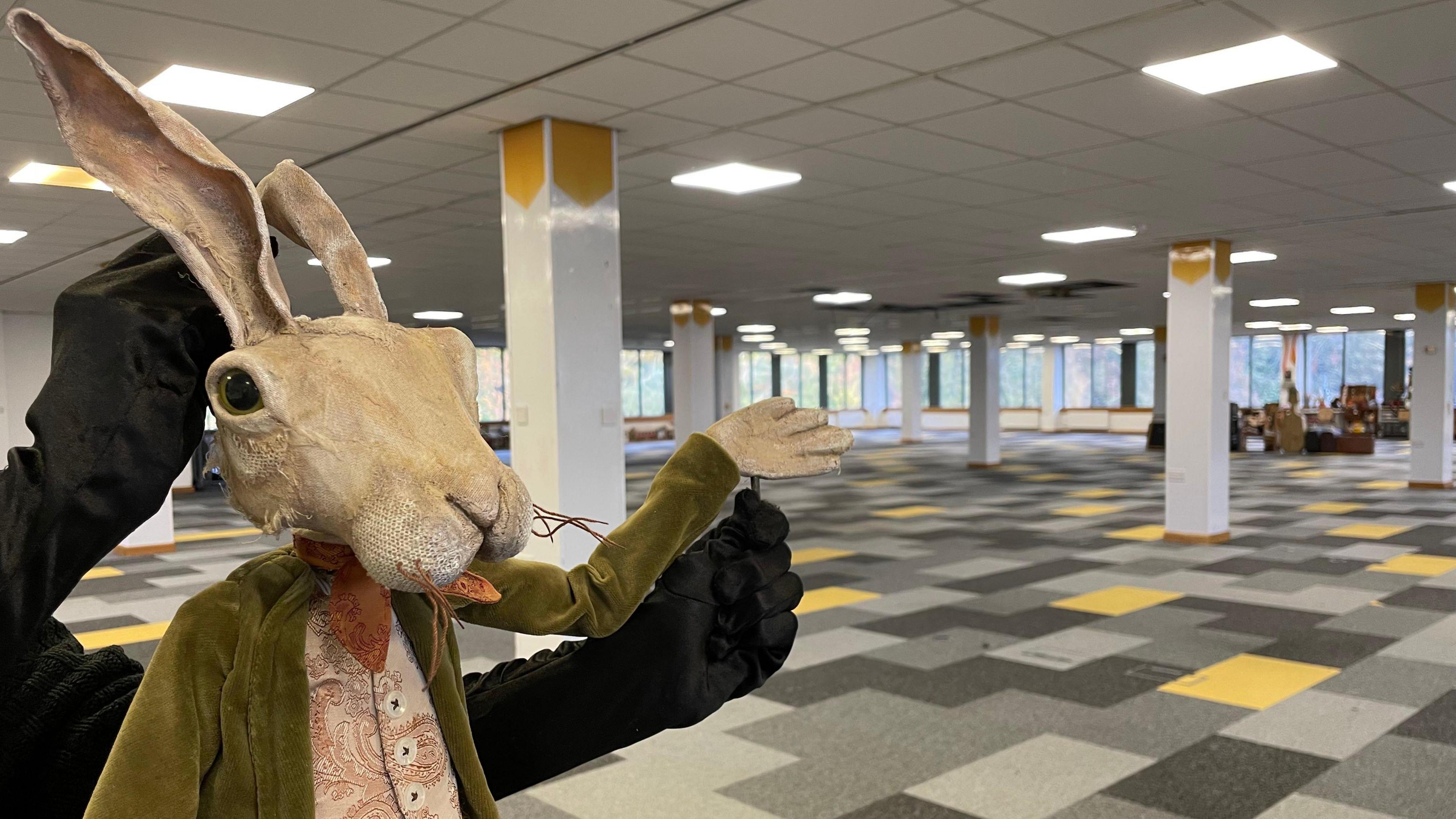 A big rabbit puppet wearing a paisley waistcoat, orange scarf and green jacket with it's arm raised signalling the room behind it. The room is large with grey and yellow carpet and pillars throughout the room. The arms of the puppeteer can be seen behind the rabbit.