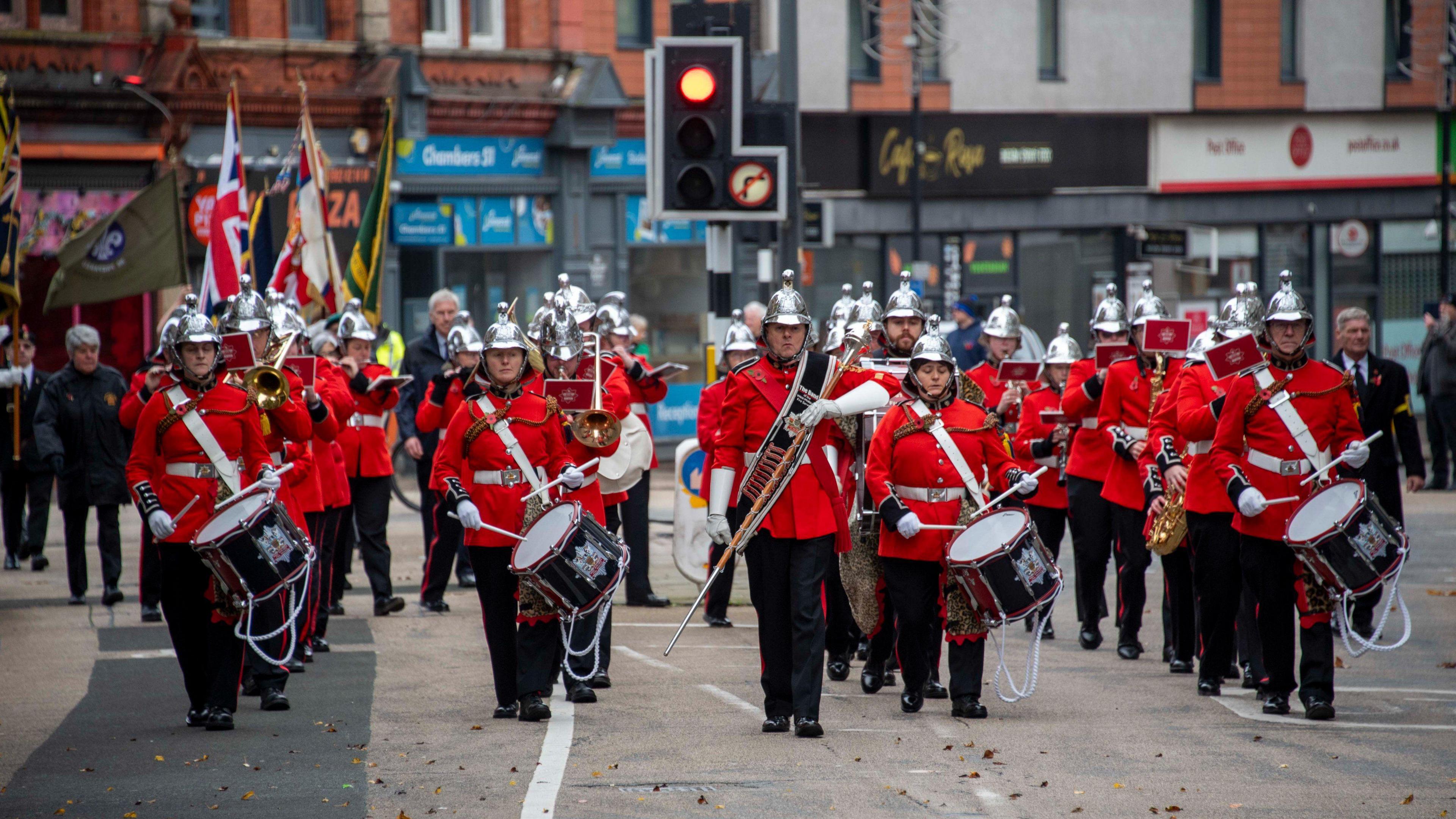 Rows of men and women in red military uniforms, with black trousers and white helmets, holding a variety of musical instruments in a street, with a row of shops behind them