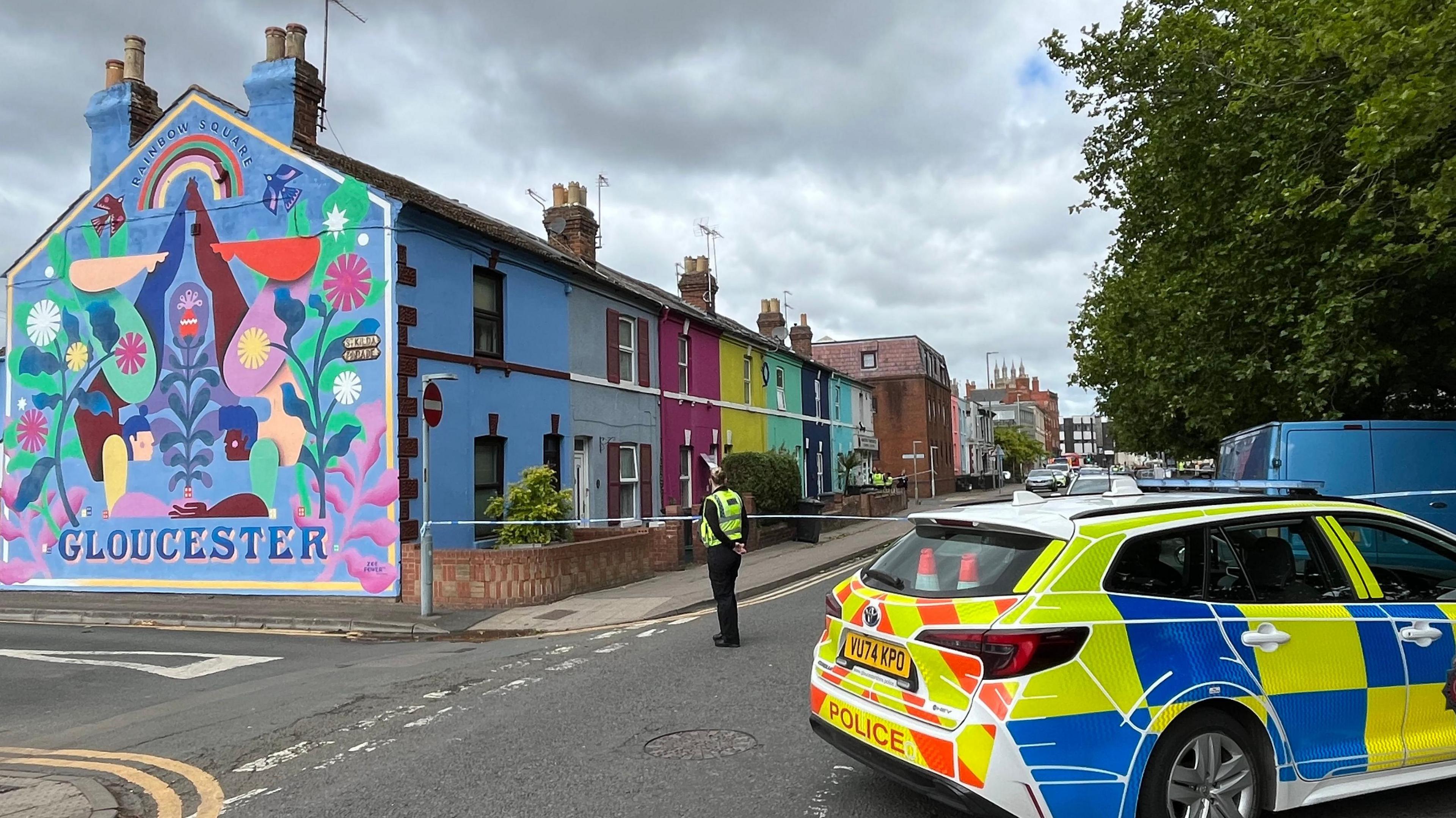 A police scene on an urban street with colourfully painted houses. A police car is parked across the road and a police officer is stood looking at the houses, with their hands behind their back. On the side of one of the houses is a colourful painted mural. There is police tape across the road. It is a cloudy day.