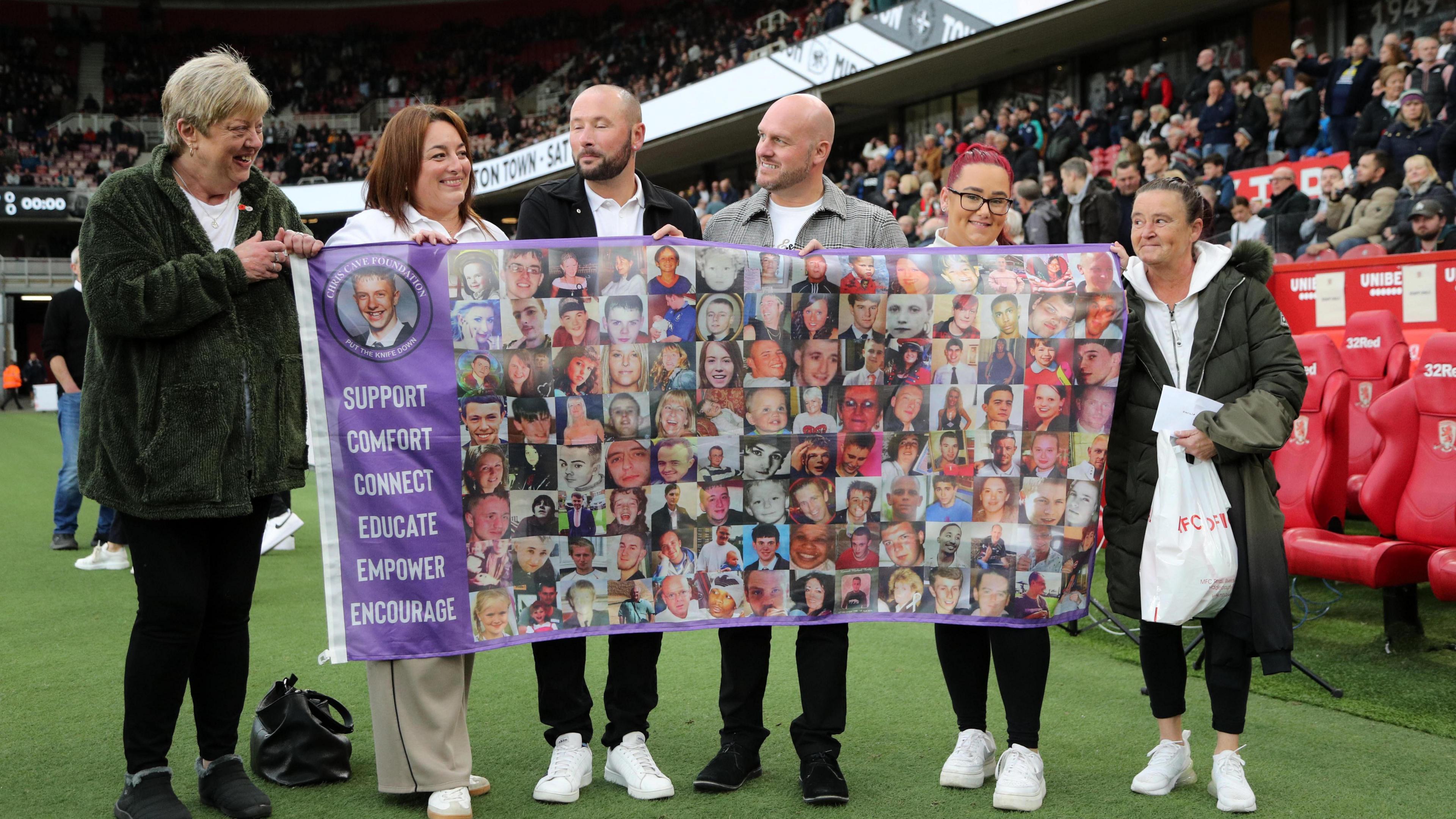 Theresa Cave and a group of people in white T-shirts stand pitch-side at the Riverside Stadium. They are holding a purple sign with the Chris Cave Foundation logo on it.