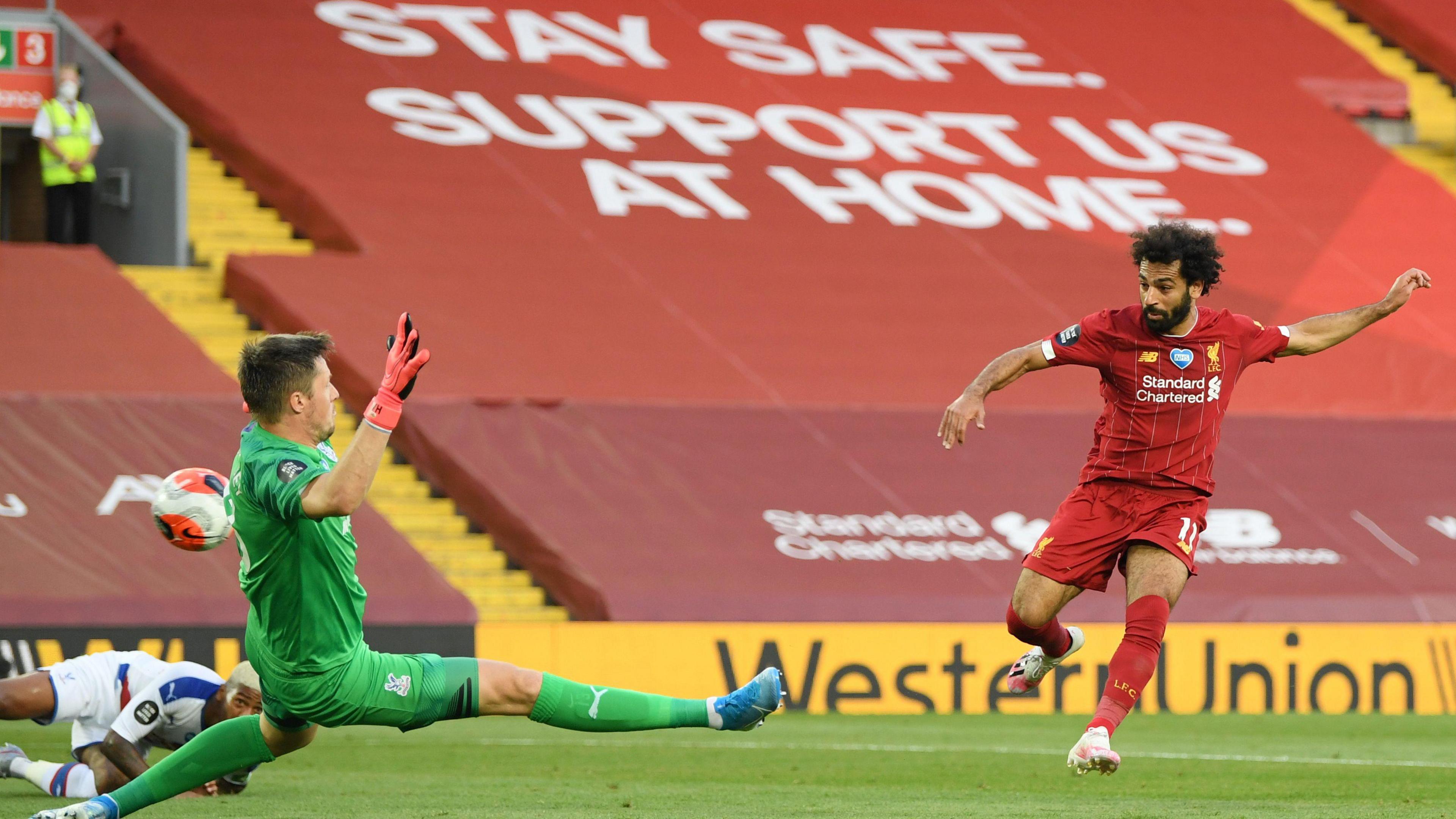 Mohamed Salah scores past the Crystal Palace goalkeeper in an empty Anfield with a large sign saying "Stay safe. Support us at home." draped over the stadium seating blocks