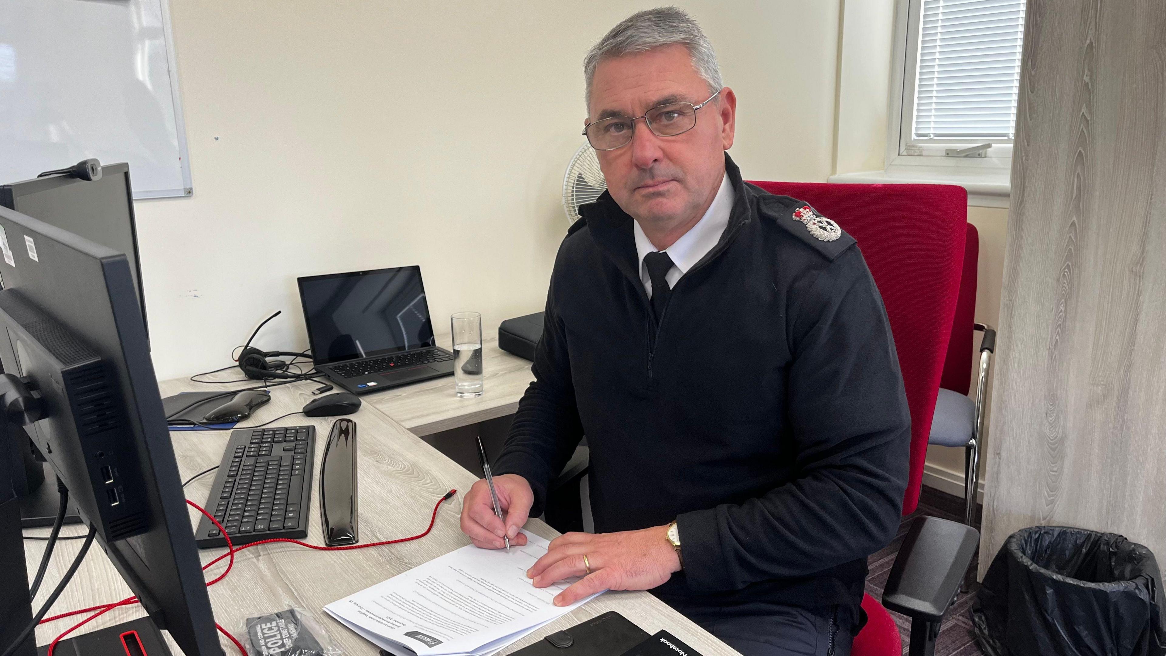 A picture of  Chief Constable James Vaughan sat at a table signing a piece of paper.