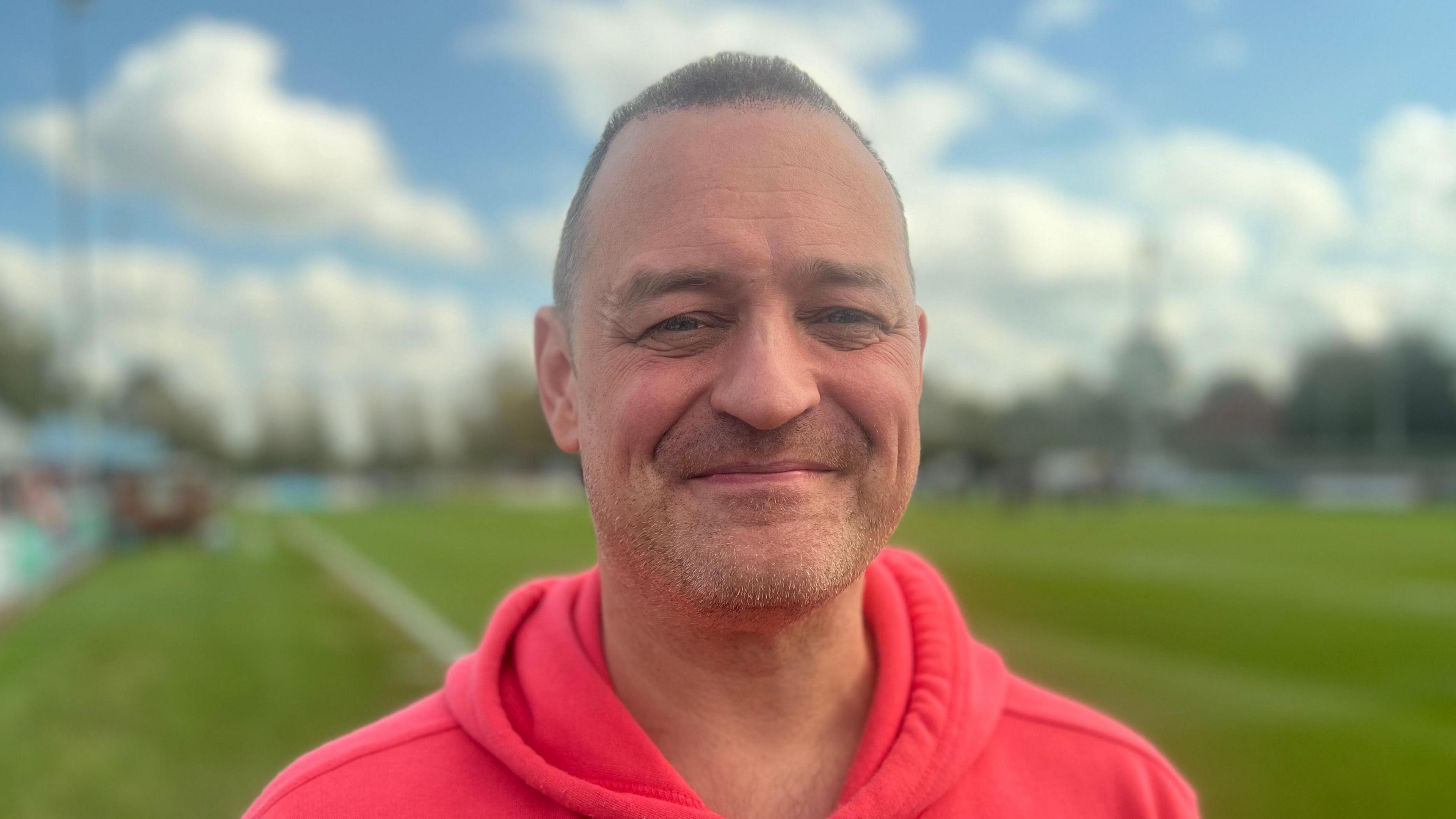 Paul Goodenough looks into the camera, wearing a bright coloured hoodie with a small smile and short hair, with a blurred football pitch in the background.