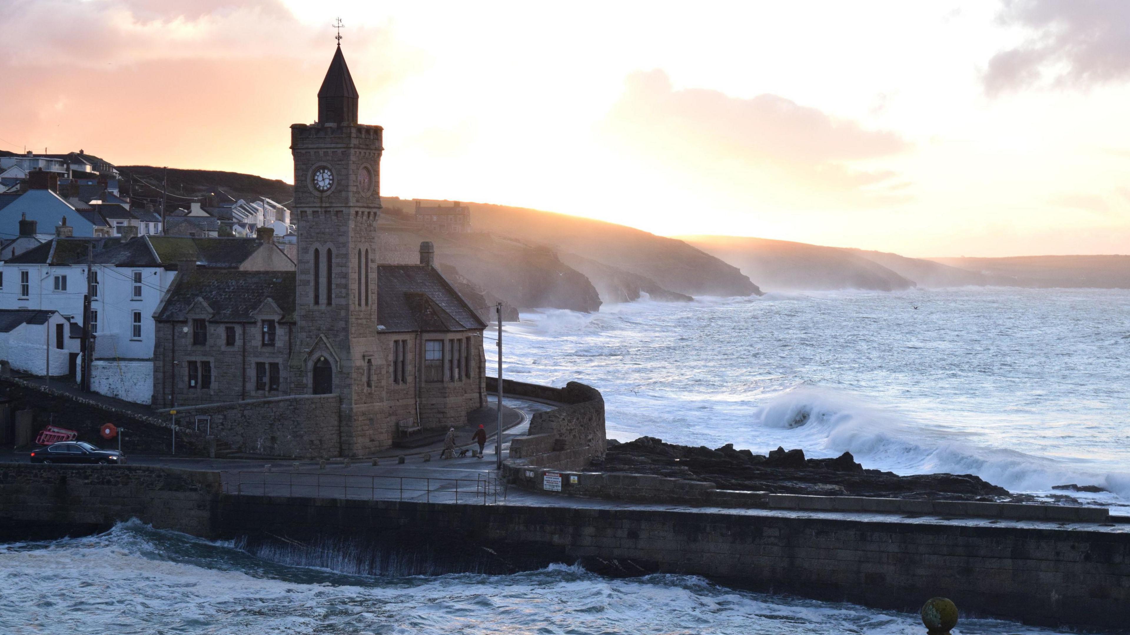 Church with tower perched next to the harbour arm in Porthleven with large waves crashing into the coast.