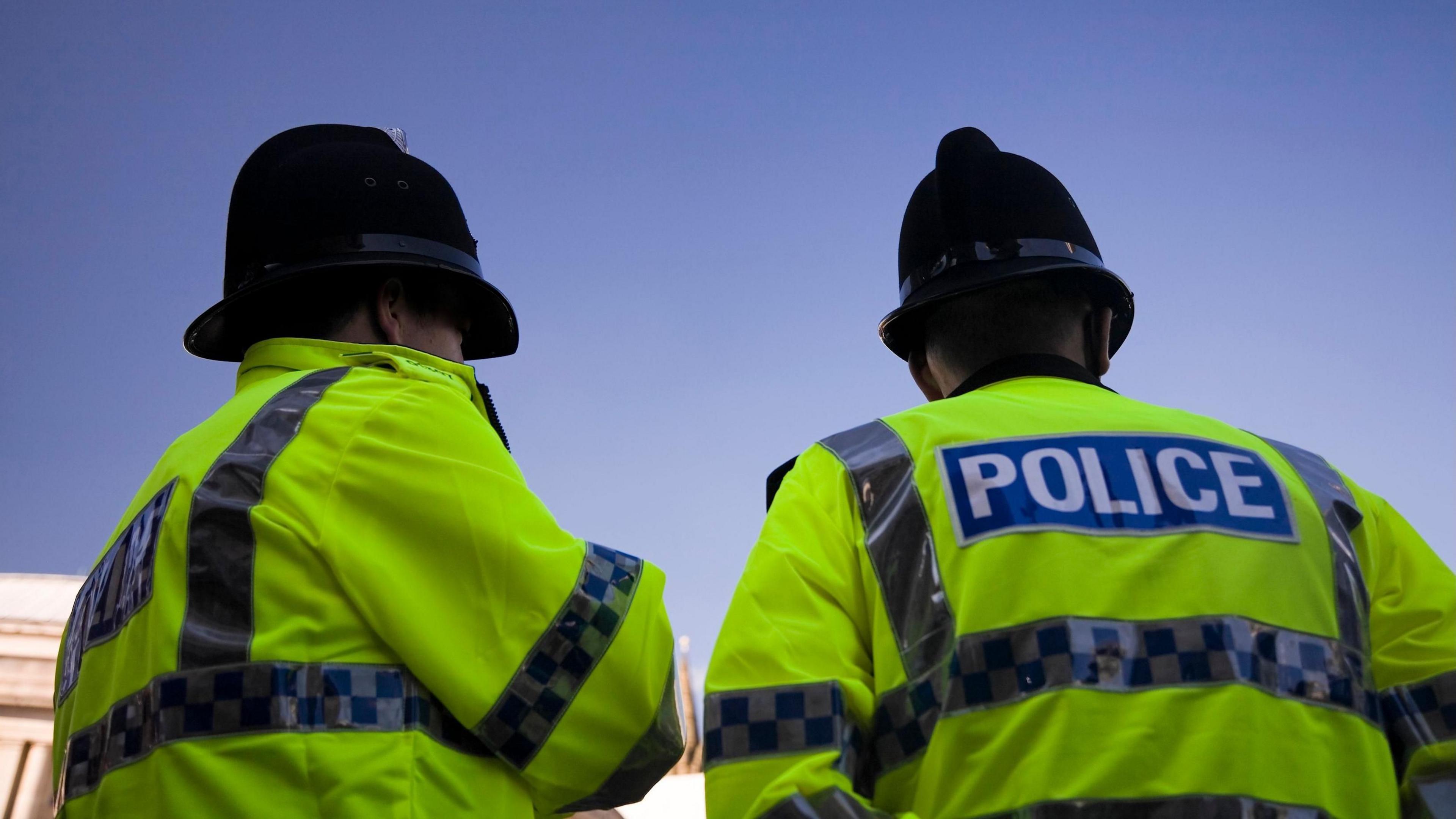 A photo from below of two police officers stood on patrol, wearing high vis police jackets and police hats. A blue sky is above them.