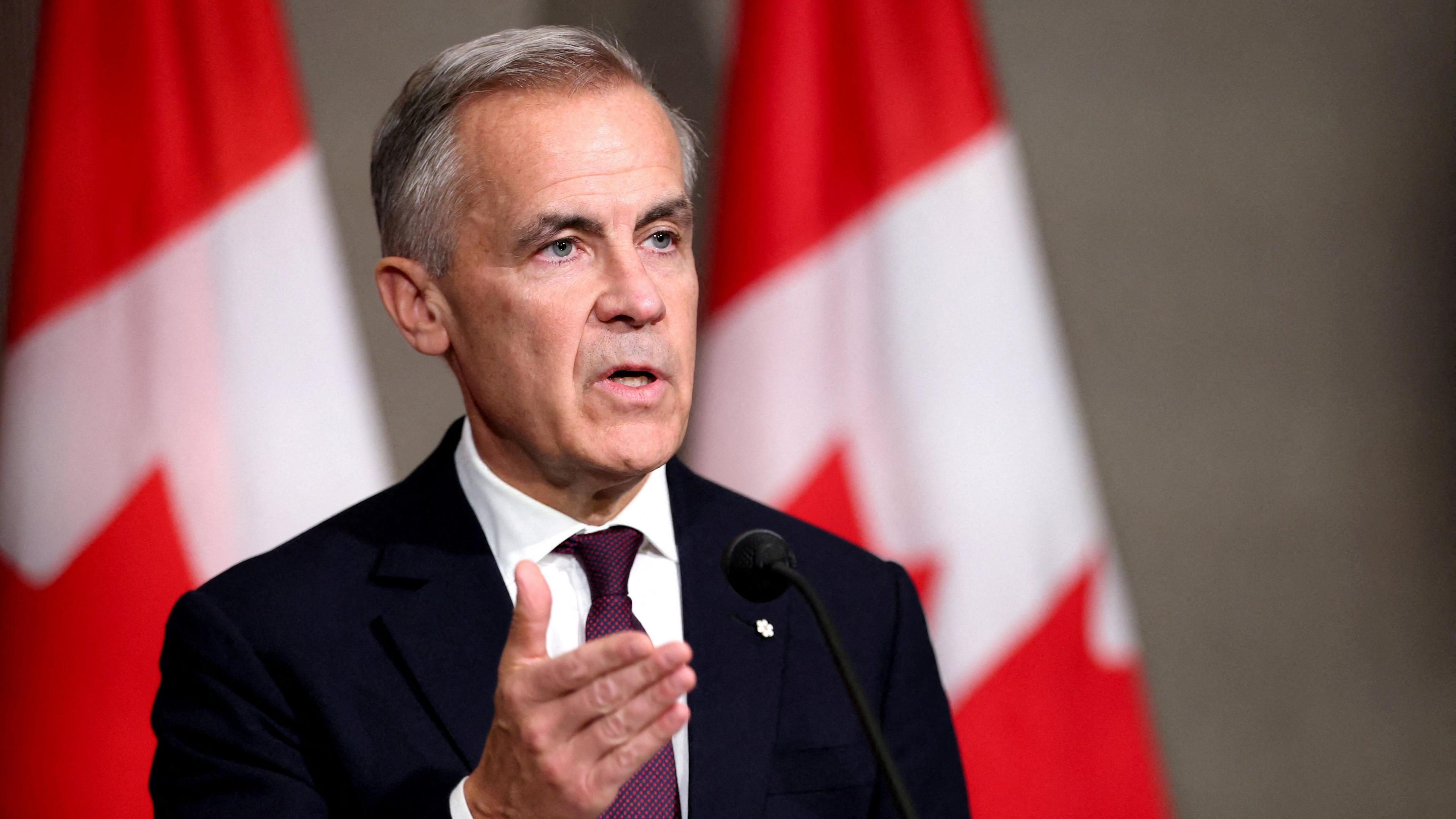 Canada's Prime Minister Mark Carney speaks during a press conference, on the sidelines of the 47th ASEAN Summit in Kuala Lumpur, Malaysia, October 27, 2025. He is speaking at a podium, wearing a black suit with a burgundy tie. Behind him are two Canadian flags. 