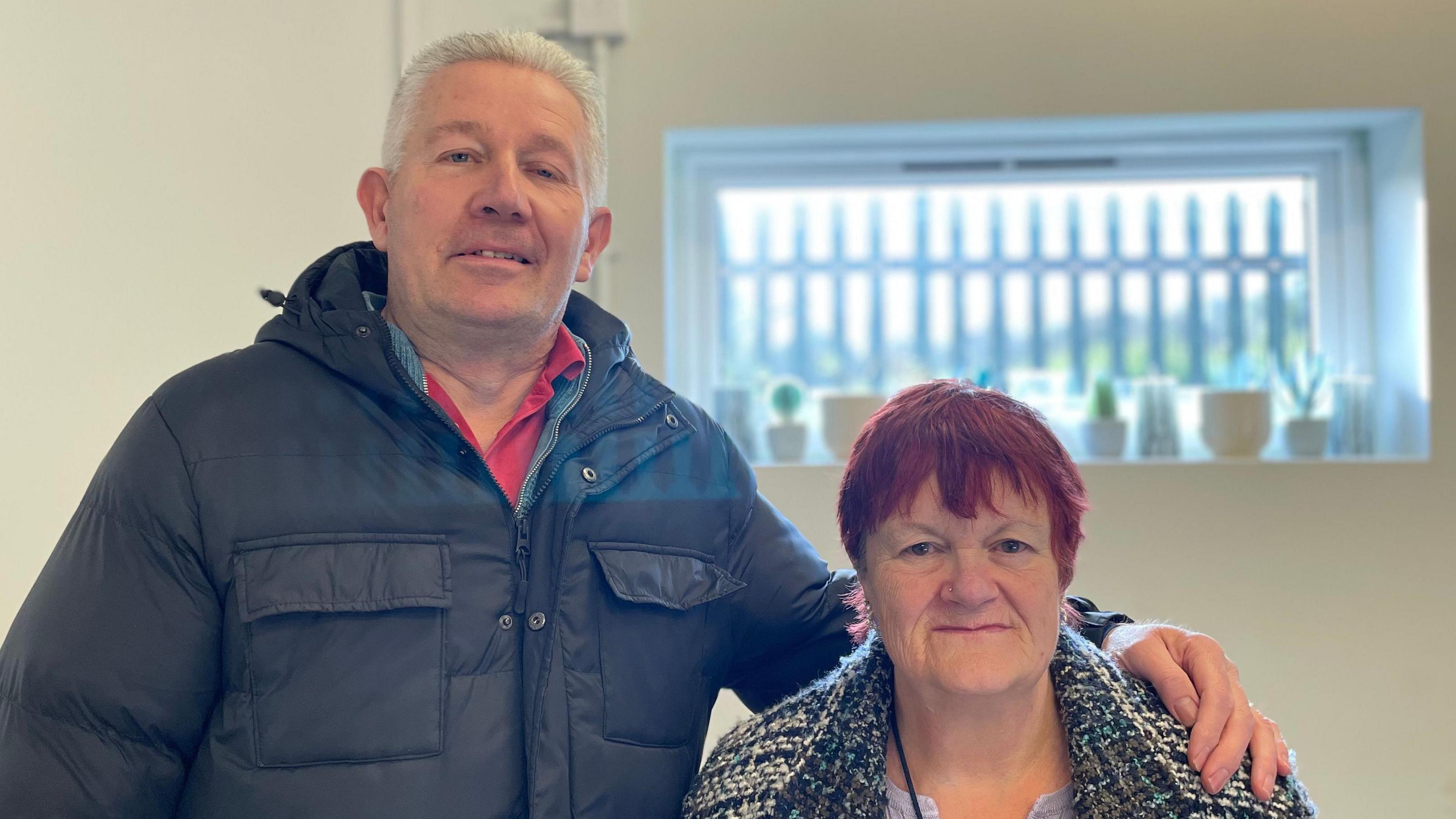 Lee is wearing a black coat and has his arm around Christina, who is wearing a black and white coat. He has short white hair and Christina has red hair.