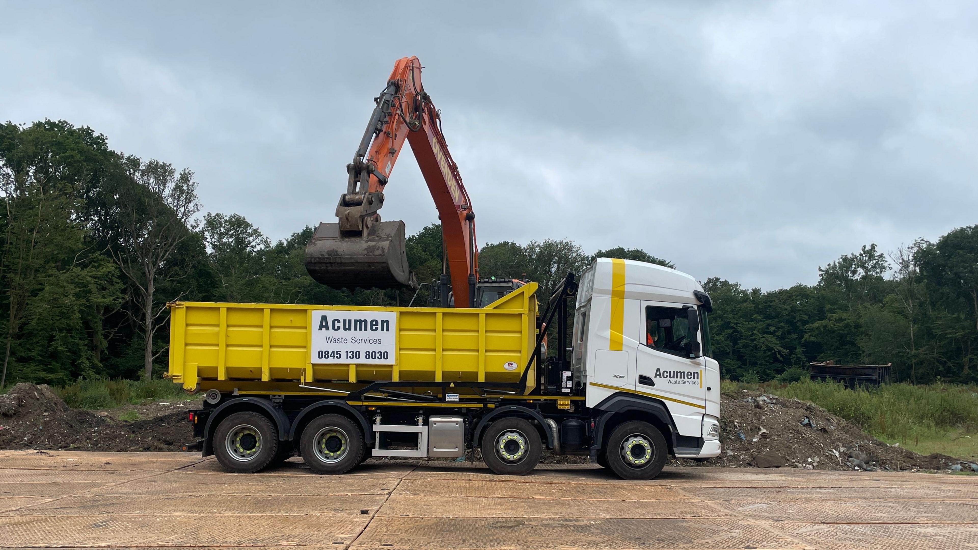 A white and yellow lorry is parked on hardstanding. A digger truck is to the side of it, with woodland behind