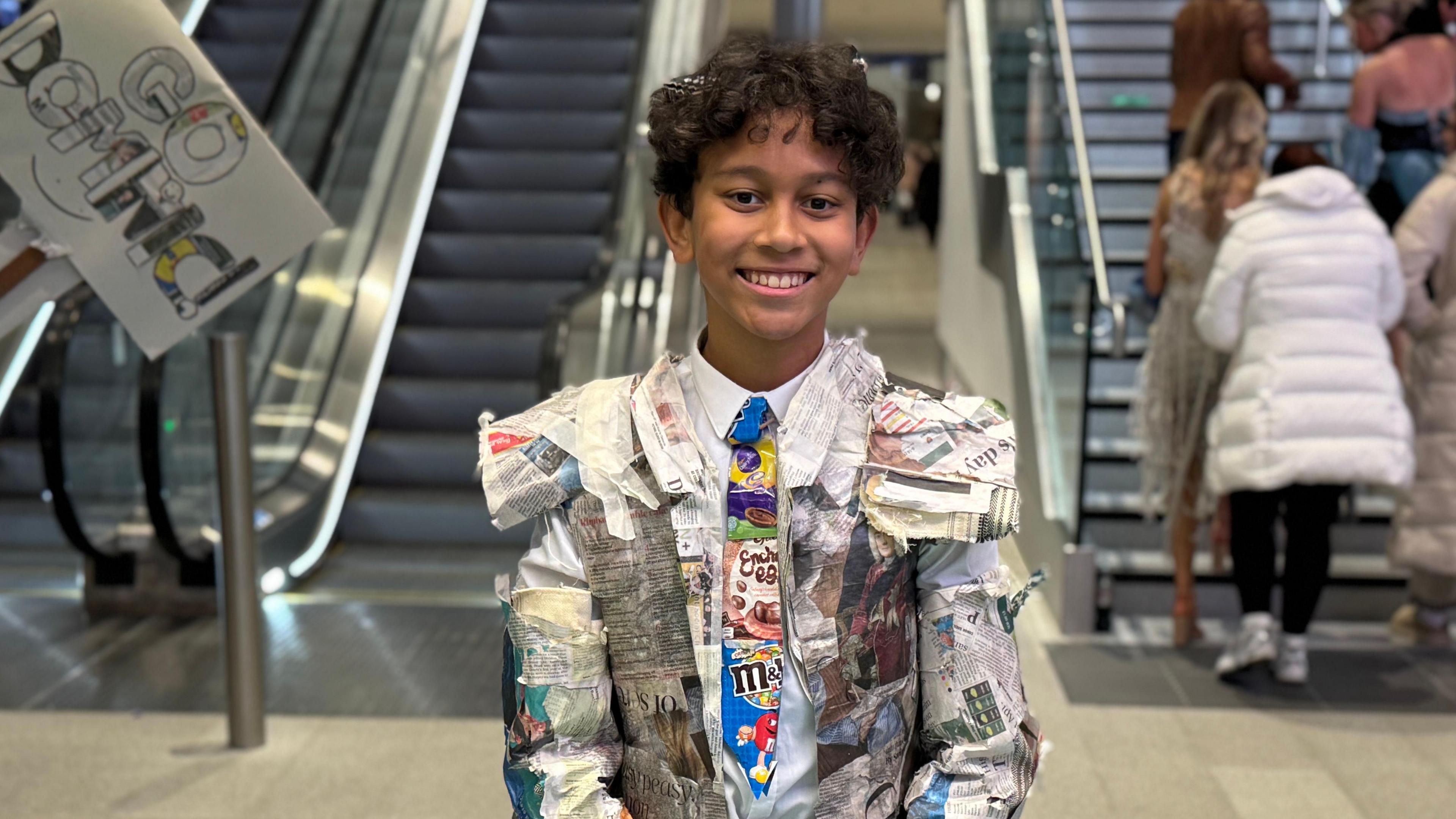Dominic wearing a suit made out of newspapers and sweet wrappers. He has short brown curly hair and is smiling at the camera. 