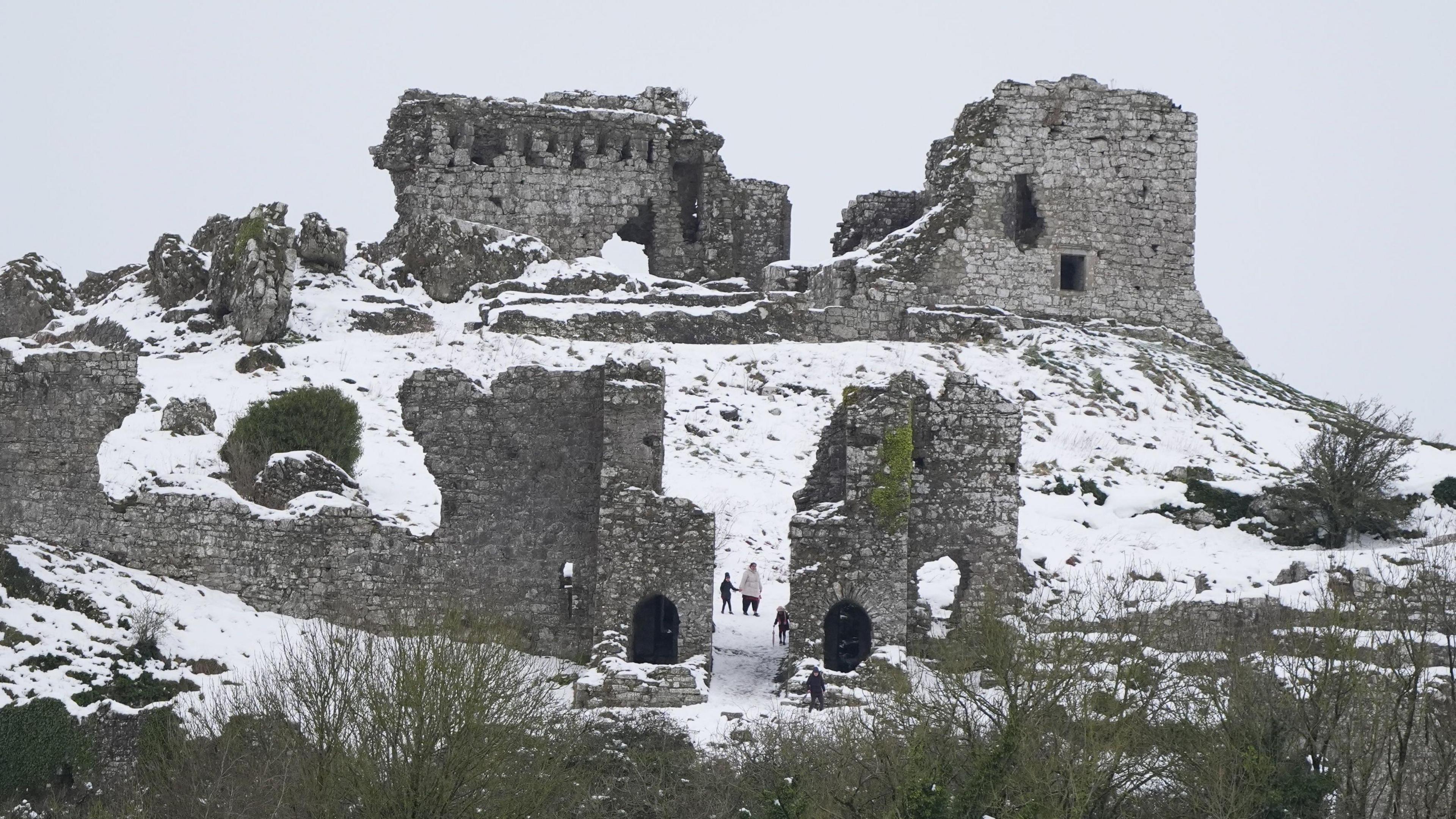 A ruined castle on a hilltop which is covered by snow. Walkers who are wrapped up warmly are walking among the ruins through the snow.