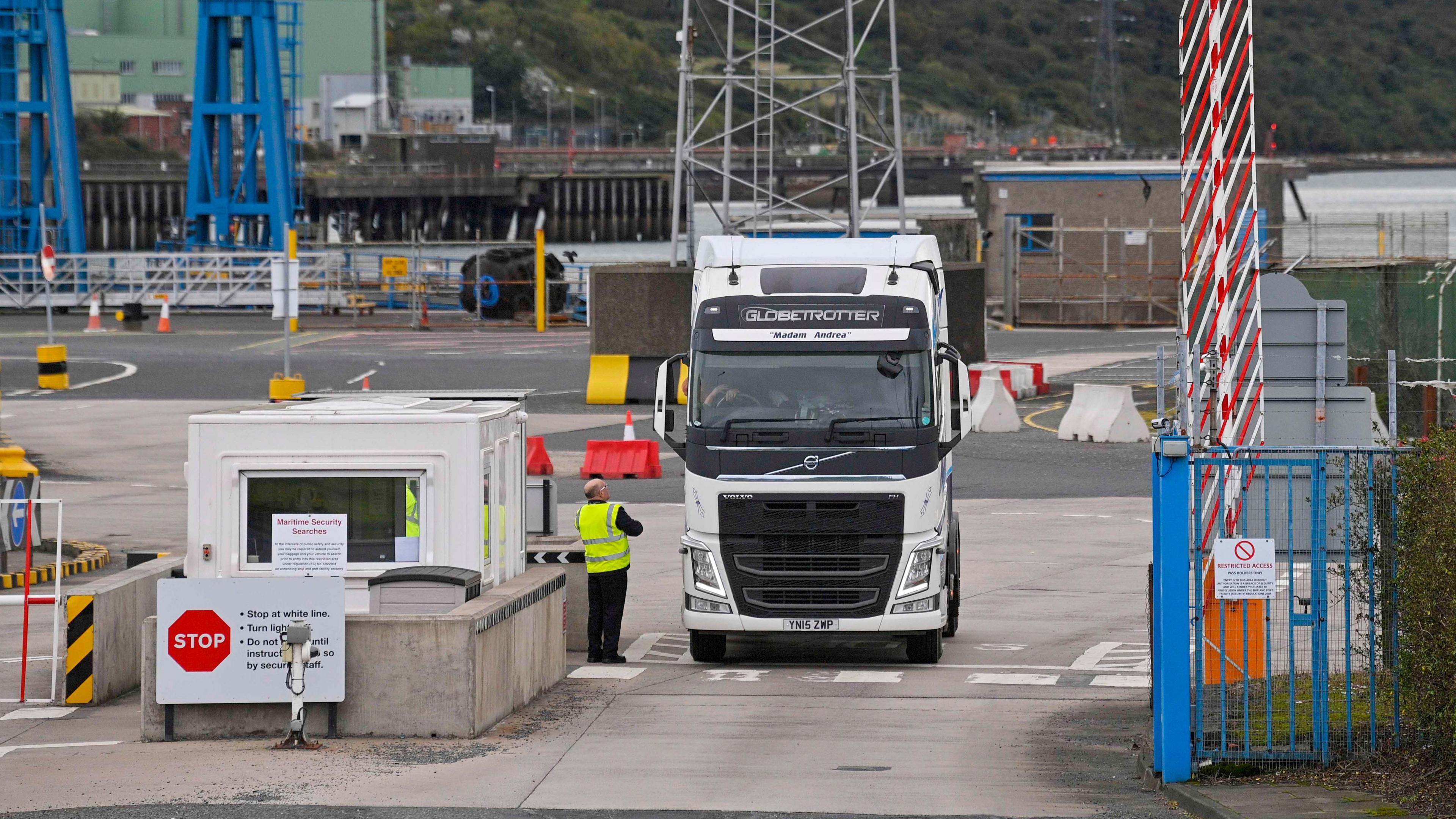 A wide shot of a white lorry leaving Larne Port. A driver is speaking to a port worker wearing a hi-vis jacket.